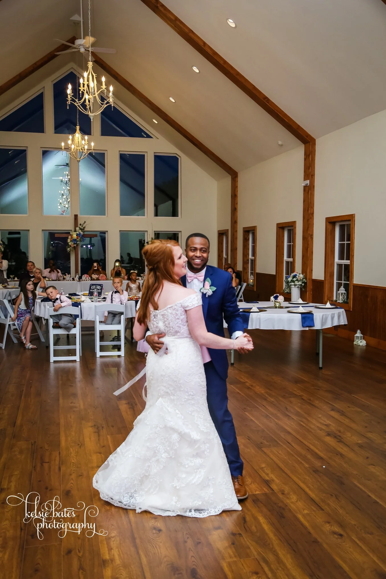 A bride and groom dancing at their wedding reception in a decorated hall with large windows and chandeliers. Guests are seated at tables in the background.