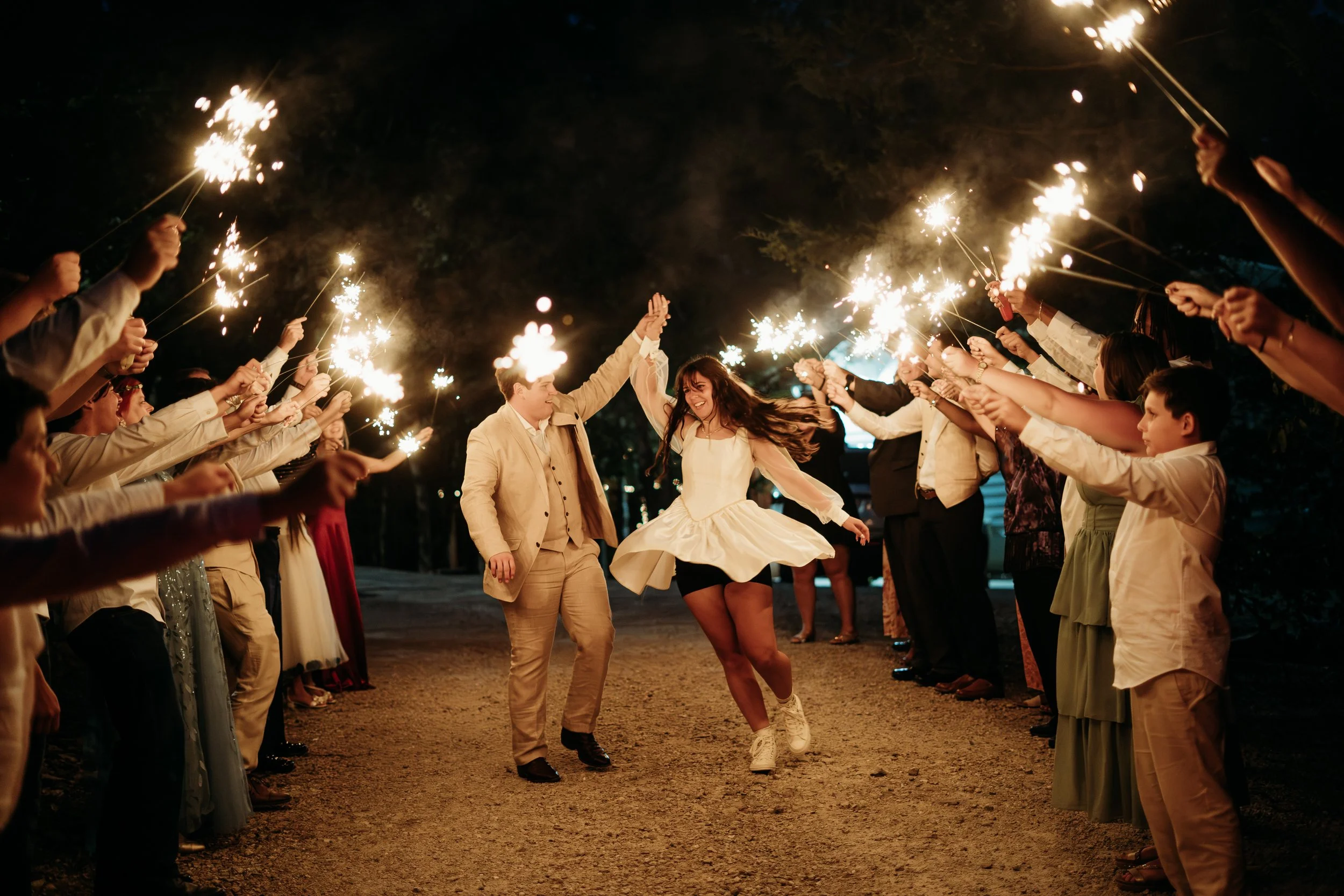 People celebrating at night with sparklers forming a circle, a couple dancing in the center, and smiles all around.