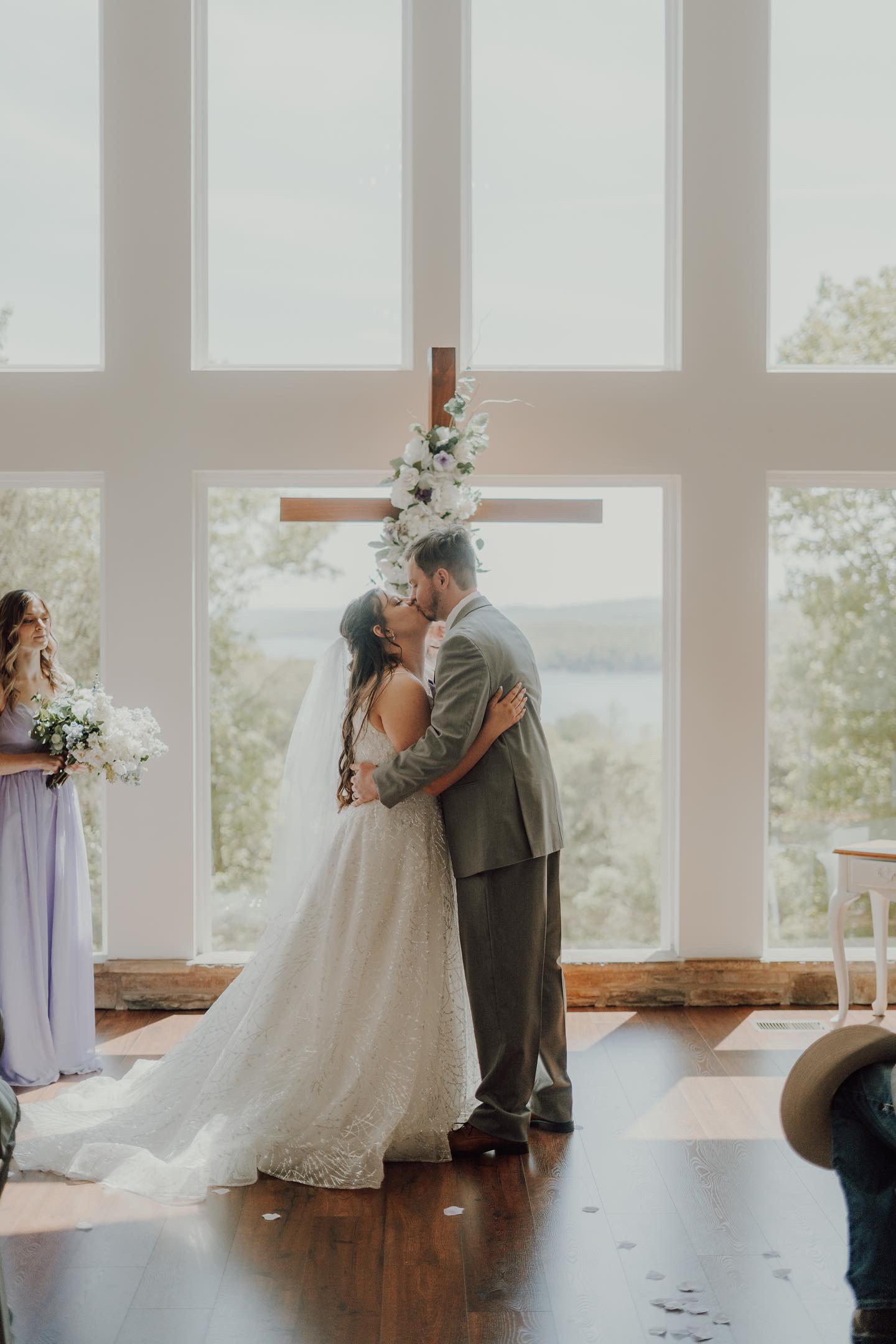 A bride and groom share a kiss during their wedding ceremony inside a church with large windows and a wooden cross decorated with white flowers behind them.