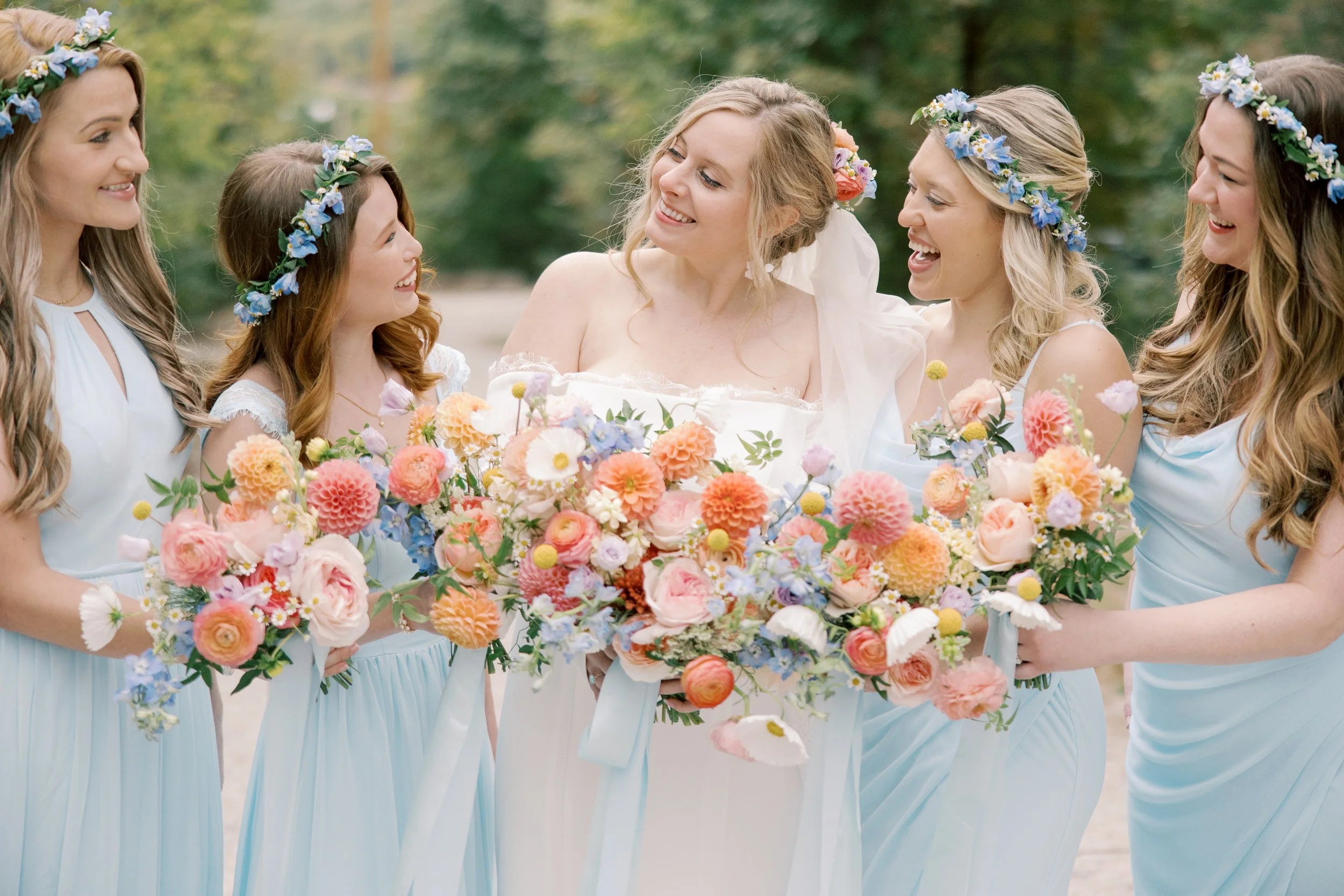 A bride and five bridesmaids wearing flower crowns and holding bouquets of colorful flowers, outdoors in a green, wooded area.