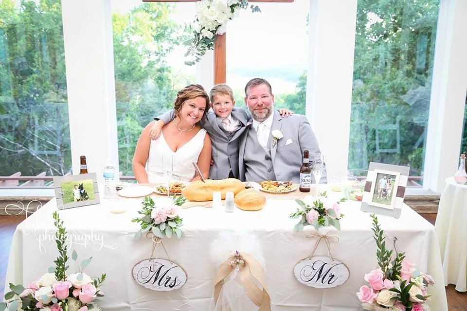 Family sitting together at a dinner table decorated with flowers and bread, in a bright room with large windows showing green trees outside. Wedding signs labeled "Mrs" and "Mr" hang on the table.
