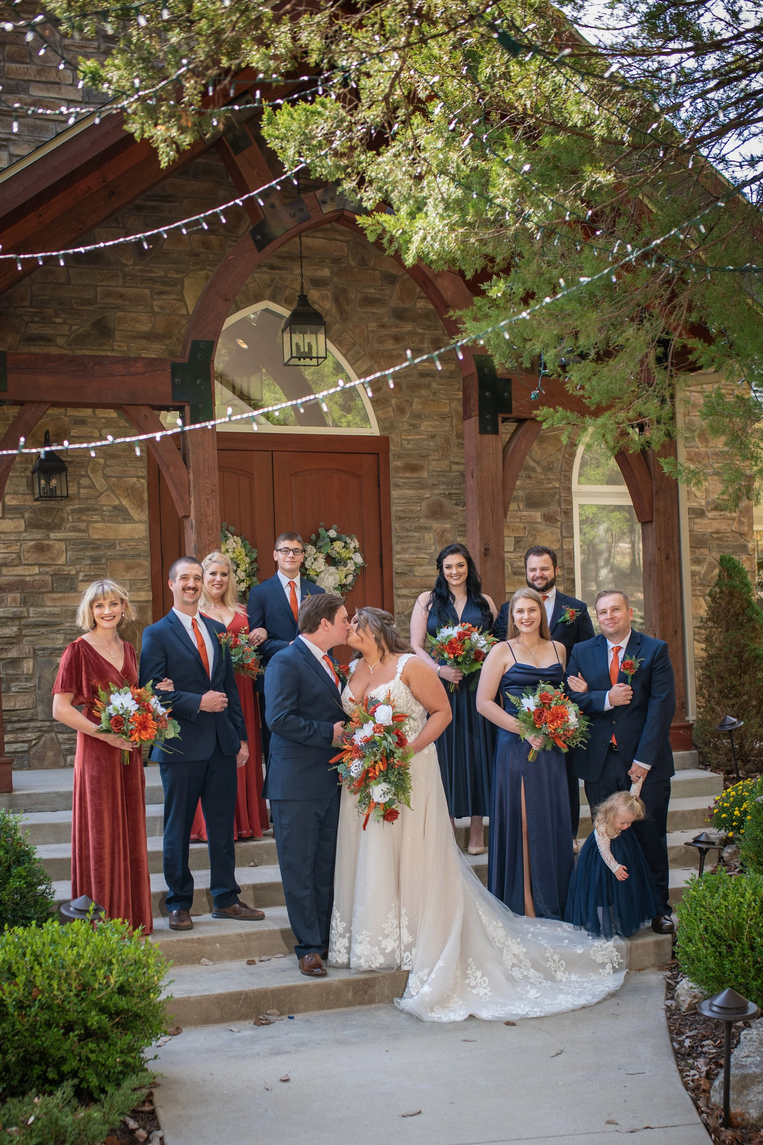 A group of people gathered on steps outside a stone house decorated for a wedding or celebration, with some holding floral bouquets, and a bride and groom in front.