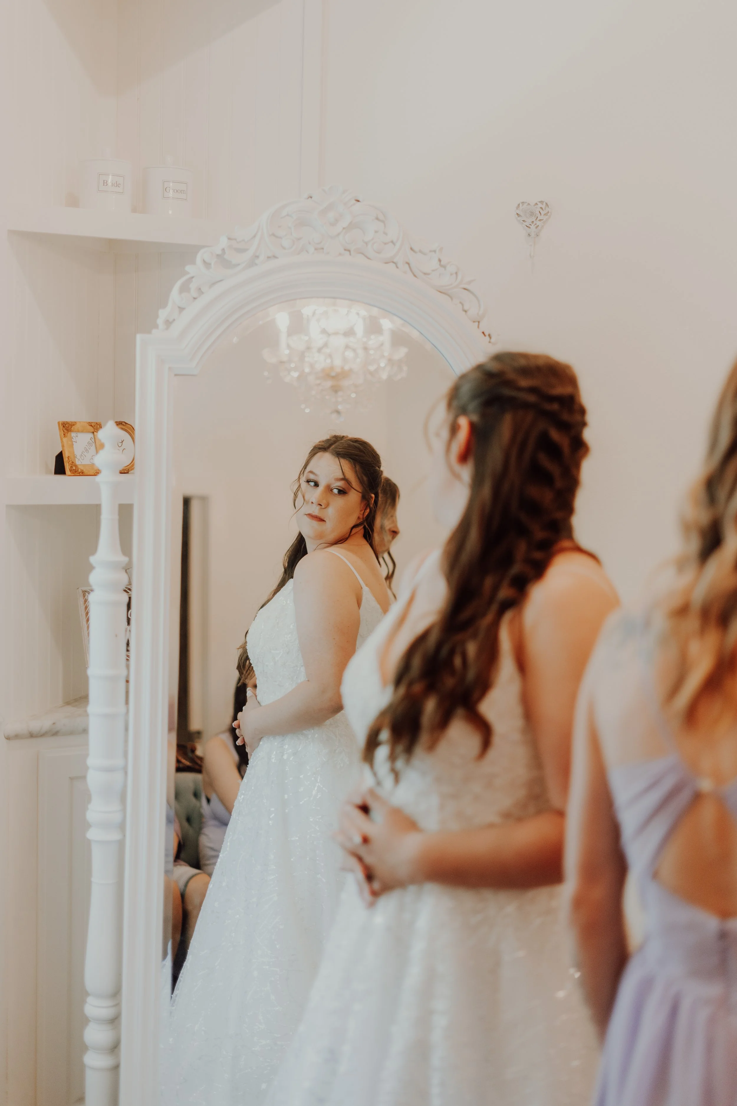 A bride in a white wedding dress looking at herself in a mirror, surrounded by women in light-colored dresses, inside a room with white walls and decorative items.