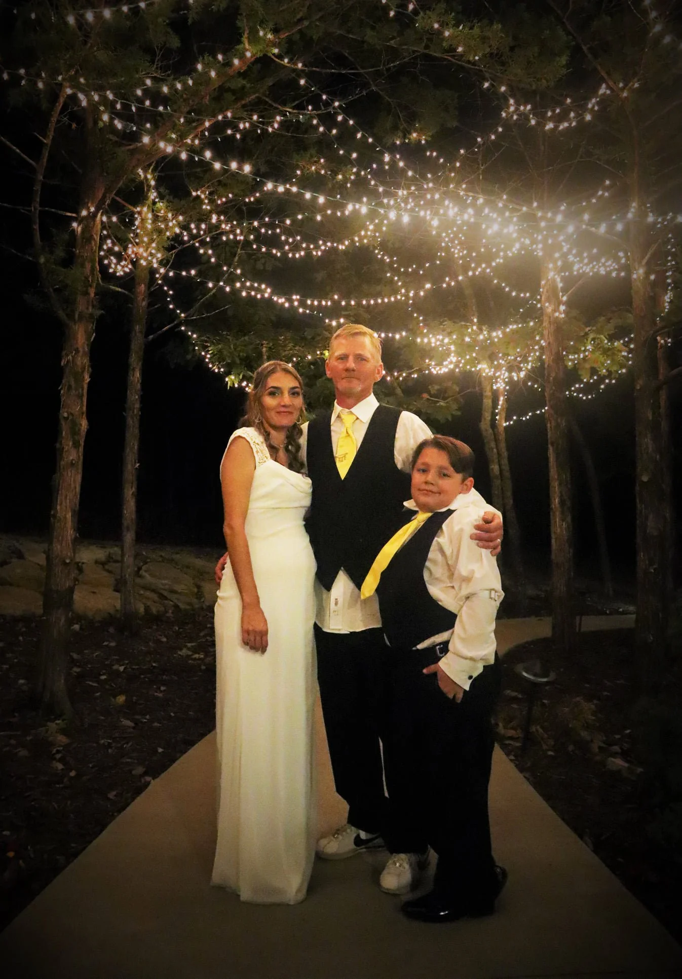 A woman in a white dress, a man in a black vest and white shirt, and a young boy in a white shirt with a yellow tie posing outdoors at night under string lights in a wooded area.
