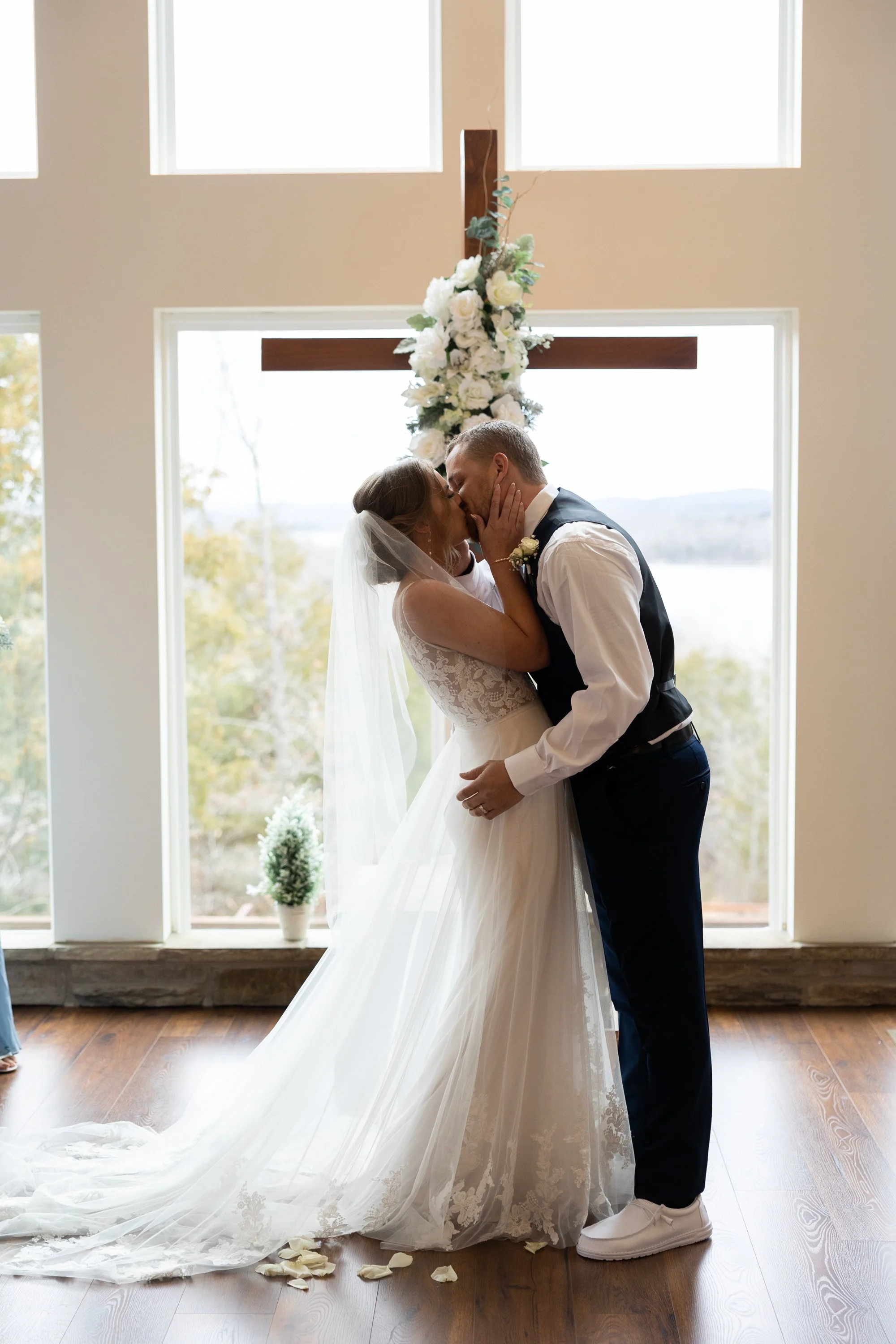 A bride and groom kissing inside a church, with the bride wearing a white wedding gown and veil, and the groom in a black vest and white shirt. Behind them is a large window with a cross and floral arrangement.