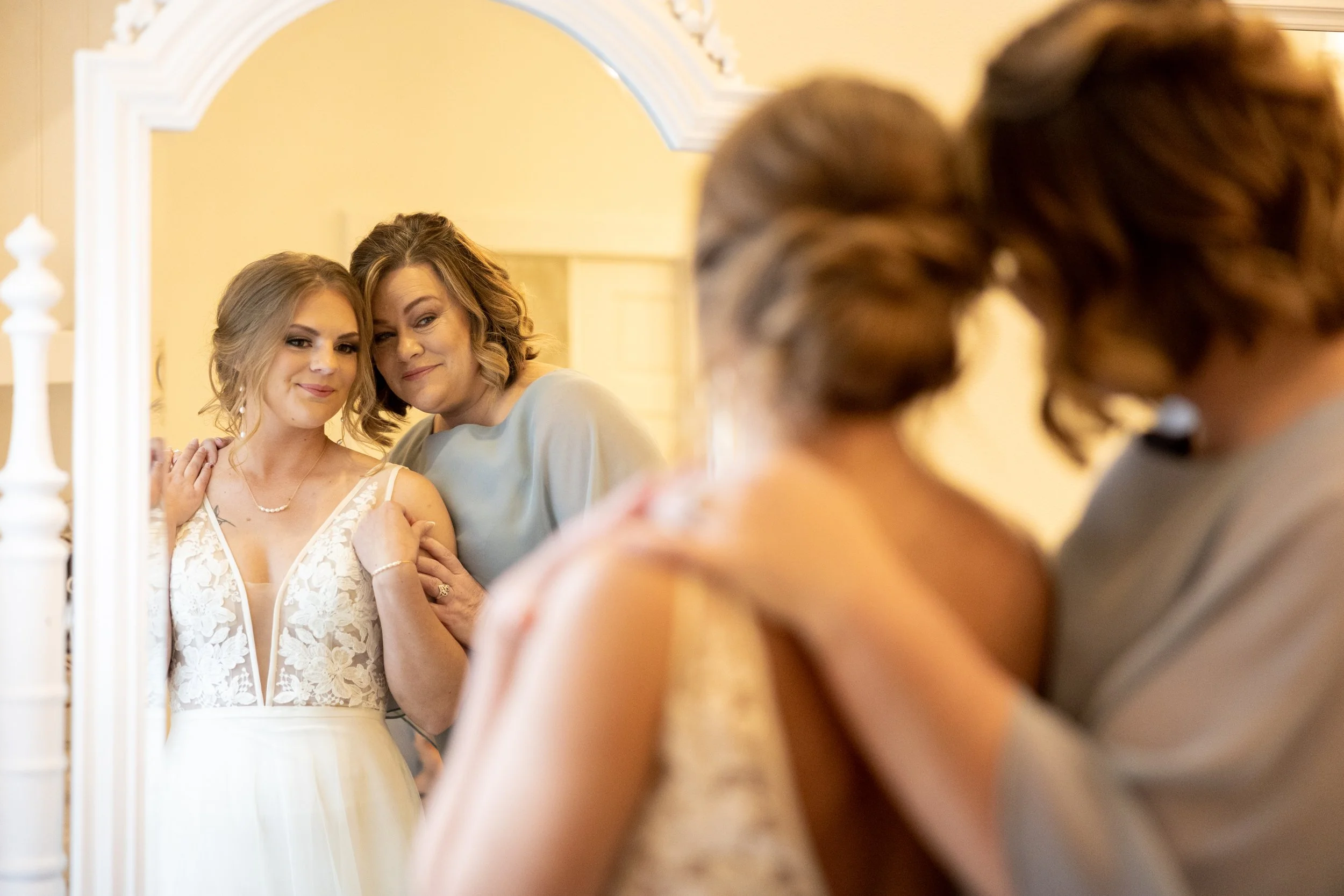Two women, one younger and one older, are looking at themselves in a mirror. The younger woman is wearing a white lace dress, and the older woman is gently holding her shoulder, both smiling softly.