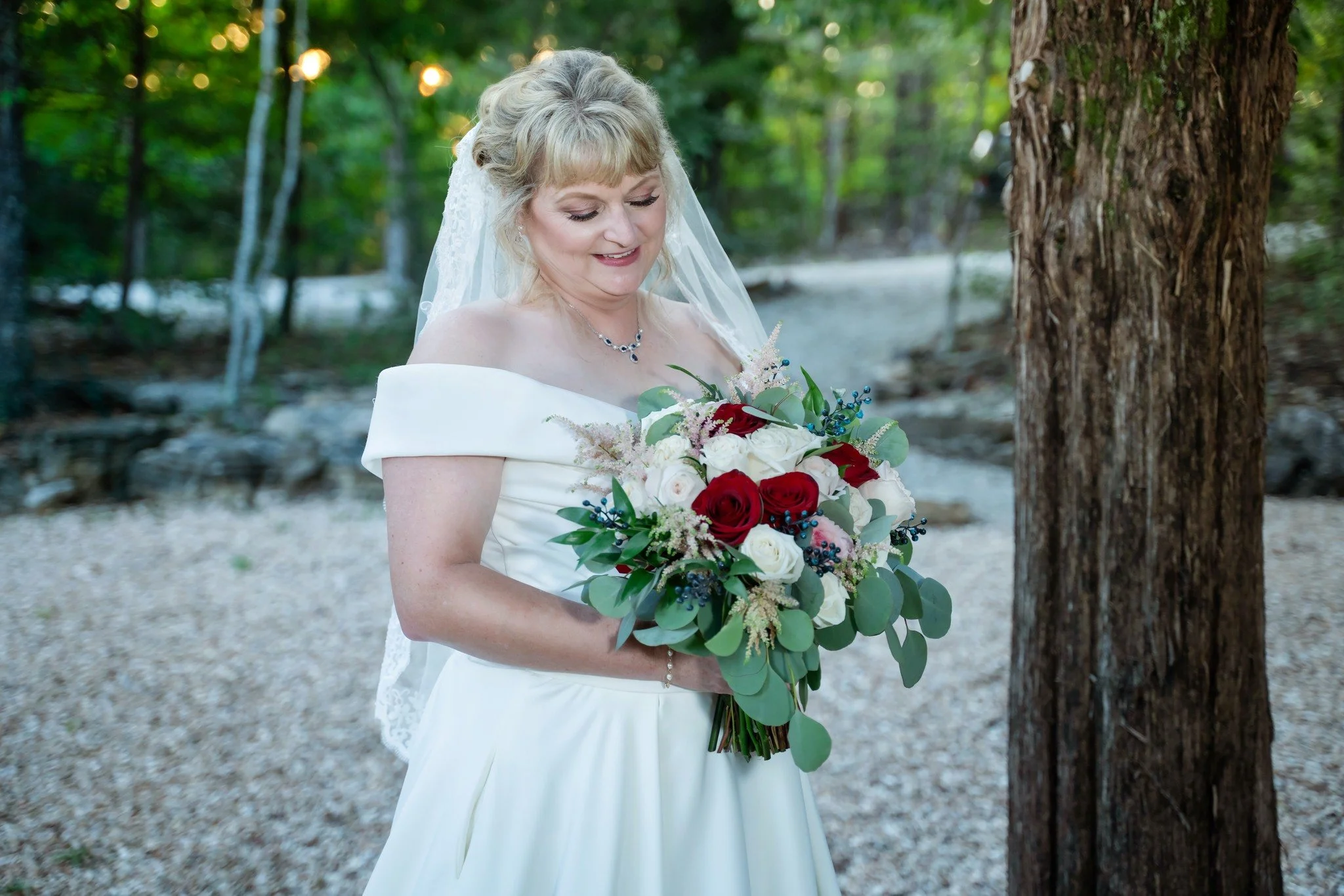 A bride in a white off-shoulder wedding dress holding a bouquet of red, white, and pink roses with greenery and blue berries in an outdoor forest setting.