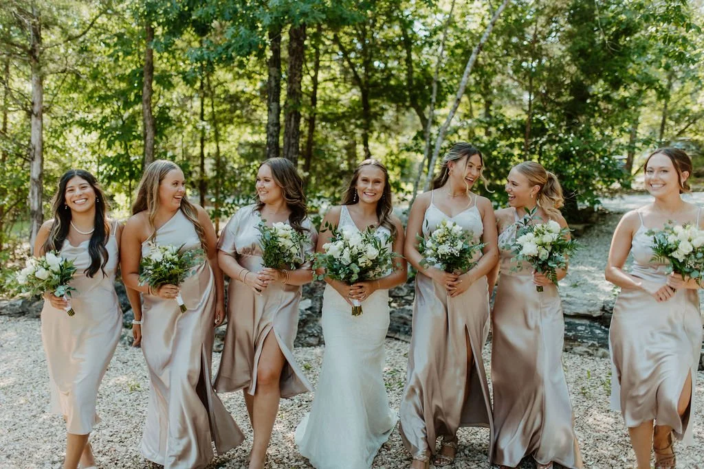 Group of seven women, including the bride in a white wedding gown, standing outdoors in a wooded area, holding bouquets of white flowers, smiling and laughing together.