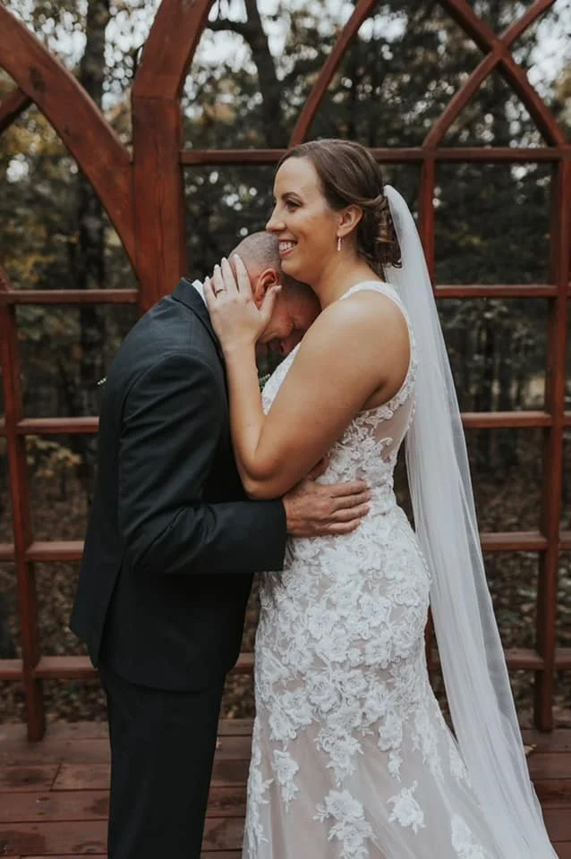 Happy bride in a white lace wedding dress and veil embracing a groom in a black suit outdoors on a wooden deck, with a wooden arch and trees in the background.