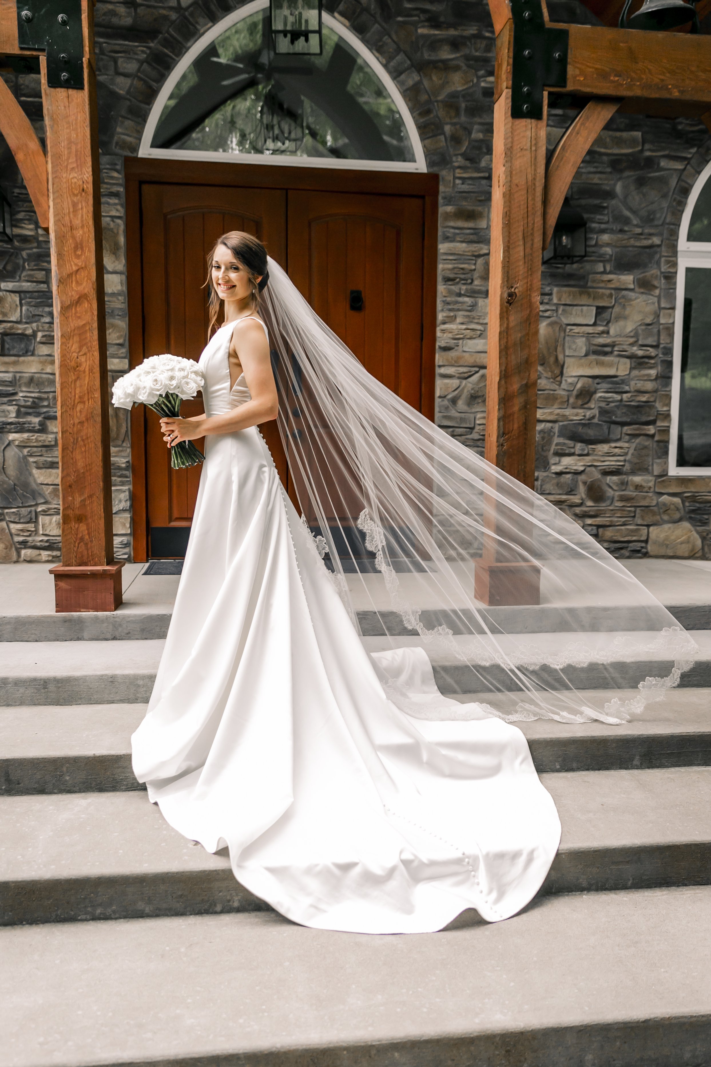 A bride in a white wedding gown holding a bouquet of white roses, standing on the steps outside a stone building with wooden accents and arched windows, smiling.