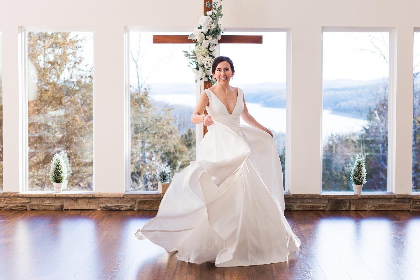 A bride in a white wedding dress dancing inside a decorated room with large windows showing an outdoor landscape with trees and water.
