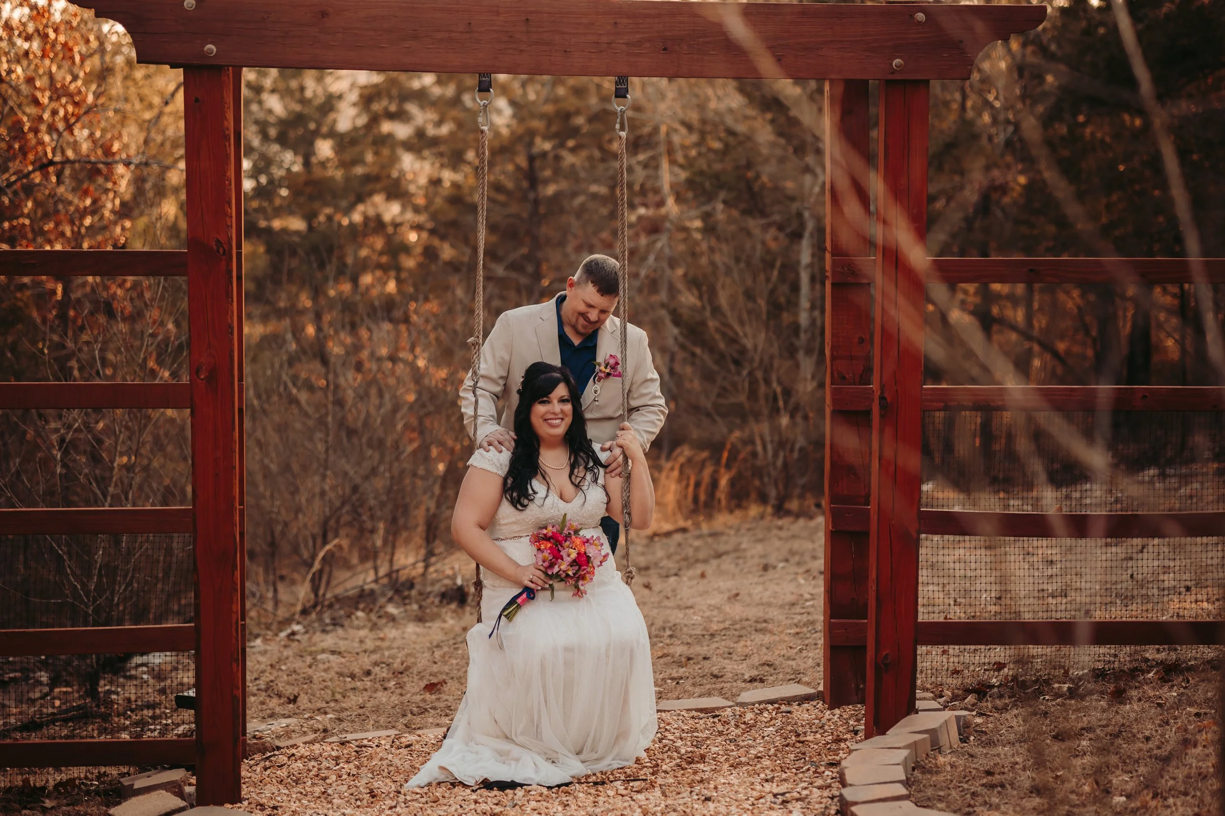 A couple at their wedding, the bride seated on a swing and holding a bouquet, the groom behind her, both smiling, outdoors during autumn with trees and fallen leaves.