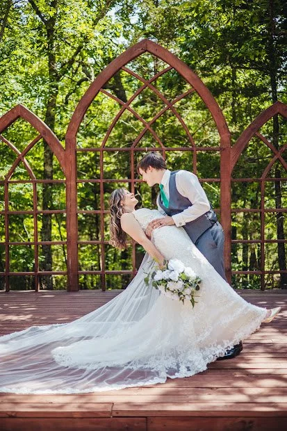A bride and groom dancing in an outdoor setting with a wooden arbor and lush green trees in the background, the bride holding a bouquet of white flowers.