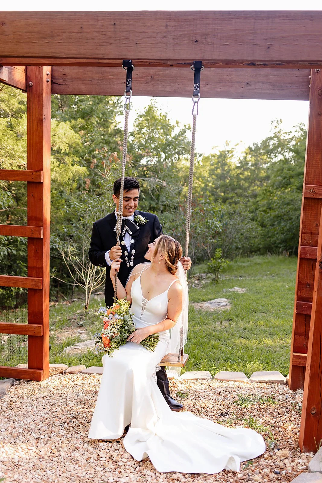 A bride and groom in wedding attire on a wooden swing outdoors, surrounded by green trees and grass, during daytime.