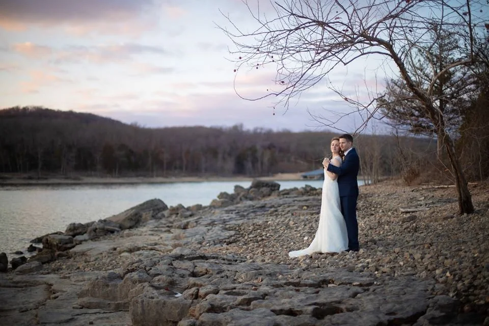 A bride and groom in wedding attire standing close together on a rocky shoreline, embracing near a leafless tree during dusk, with a river and forested hills in the background.