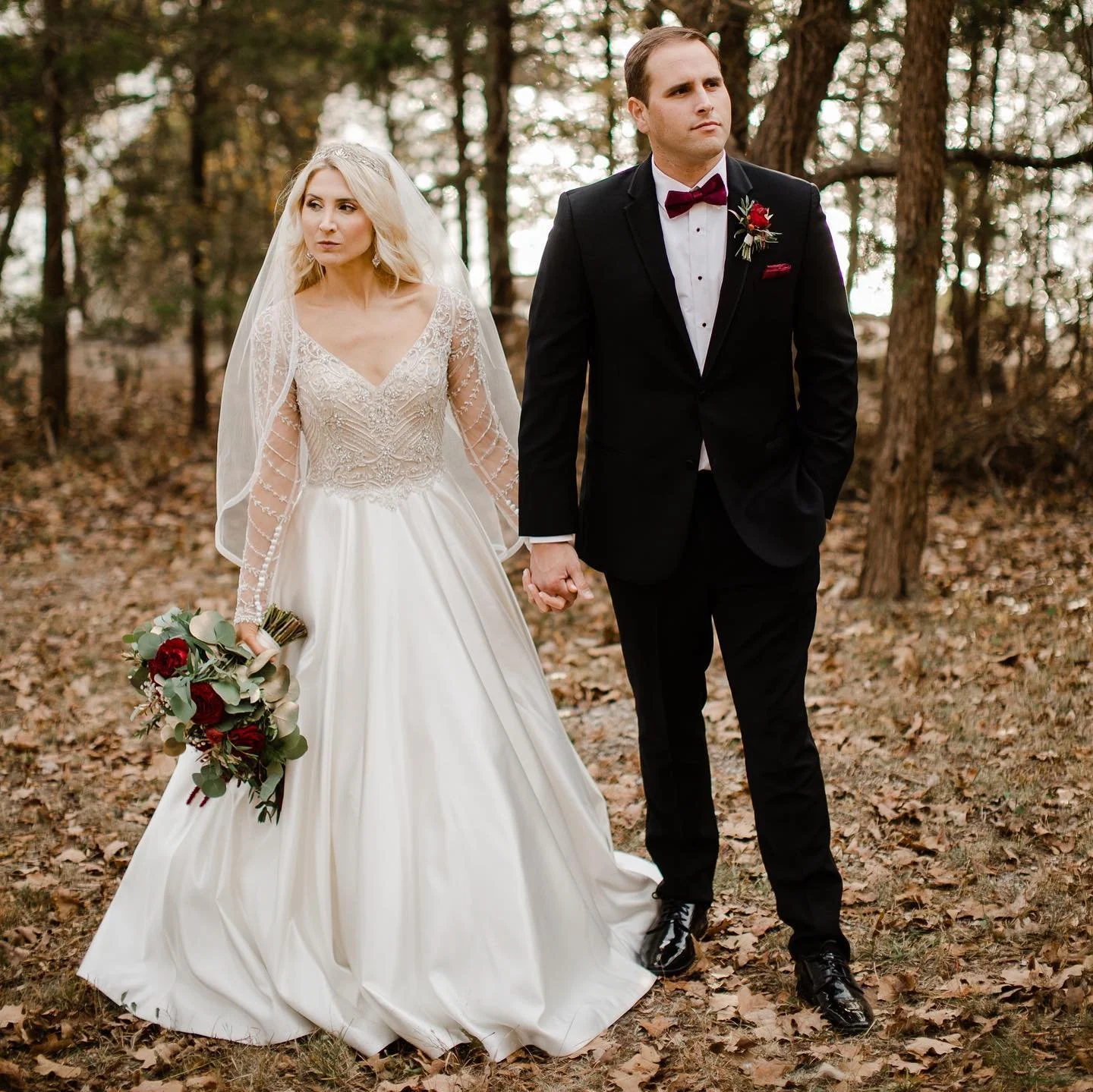 A bride and groom holding hands while walking in a wooded area during autumn. The bride wears a white wedding gown with lace details and a veil, holding a bouquet of red roses and greenery. The groom is dressed in a black tuxedo with a burgundy bow t