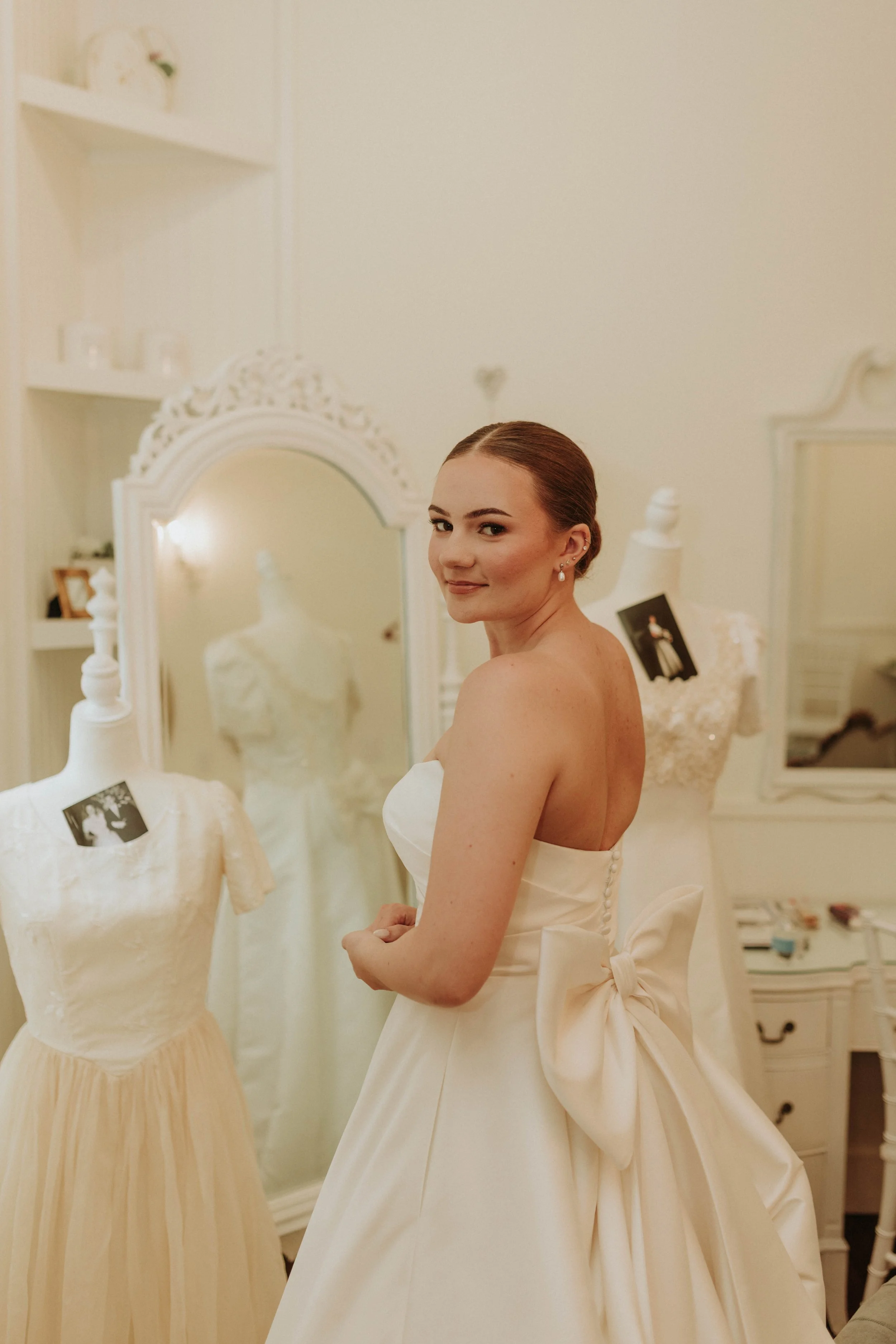 A woman with red hair in a wedding dress standing in a boutique surrounded by wedding gowns and mirrors.