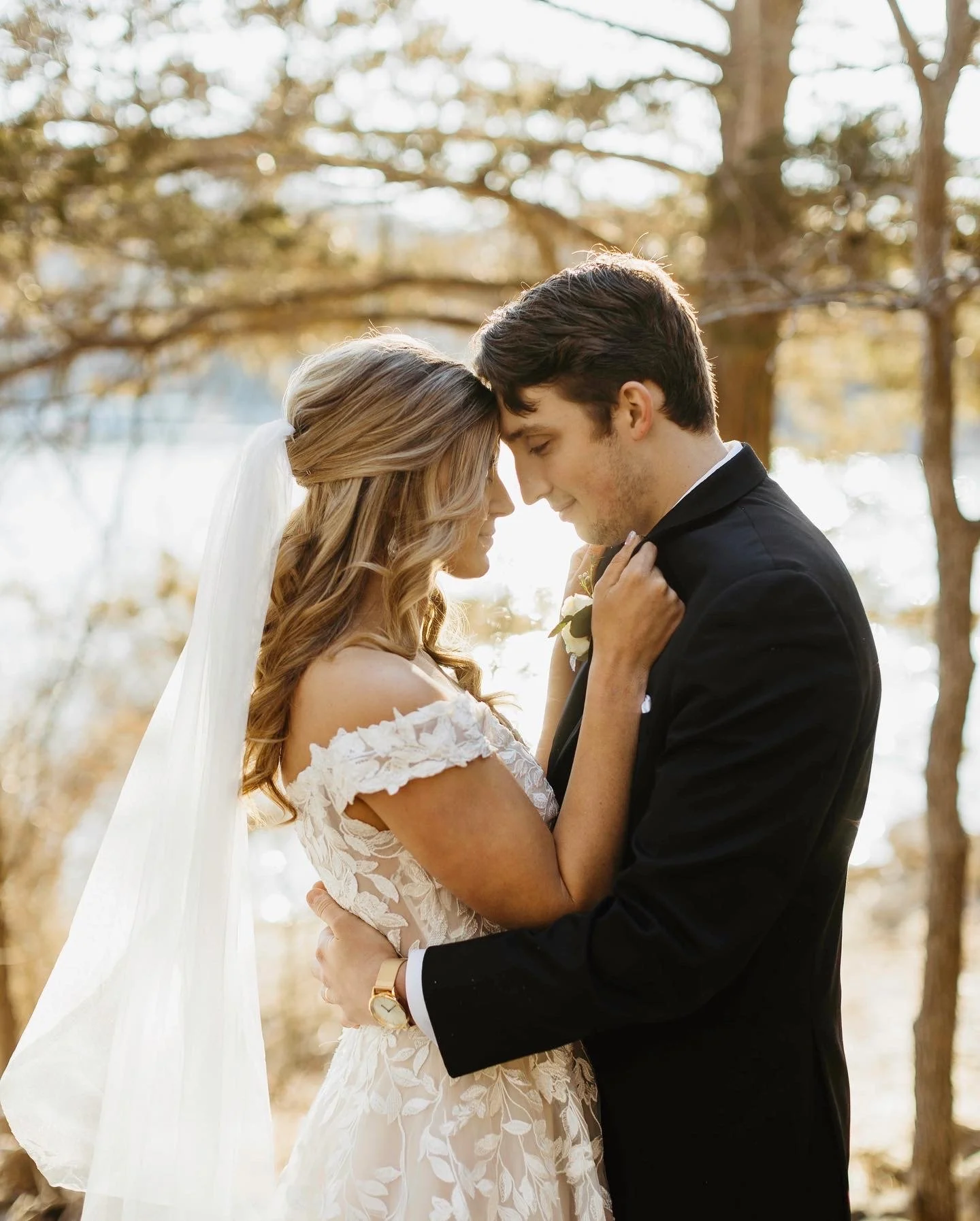 A bride and groom sharing a tender moment outdoors, standing close with foreheads touching, surrounded by trees with autumn foliage.