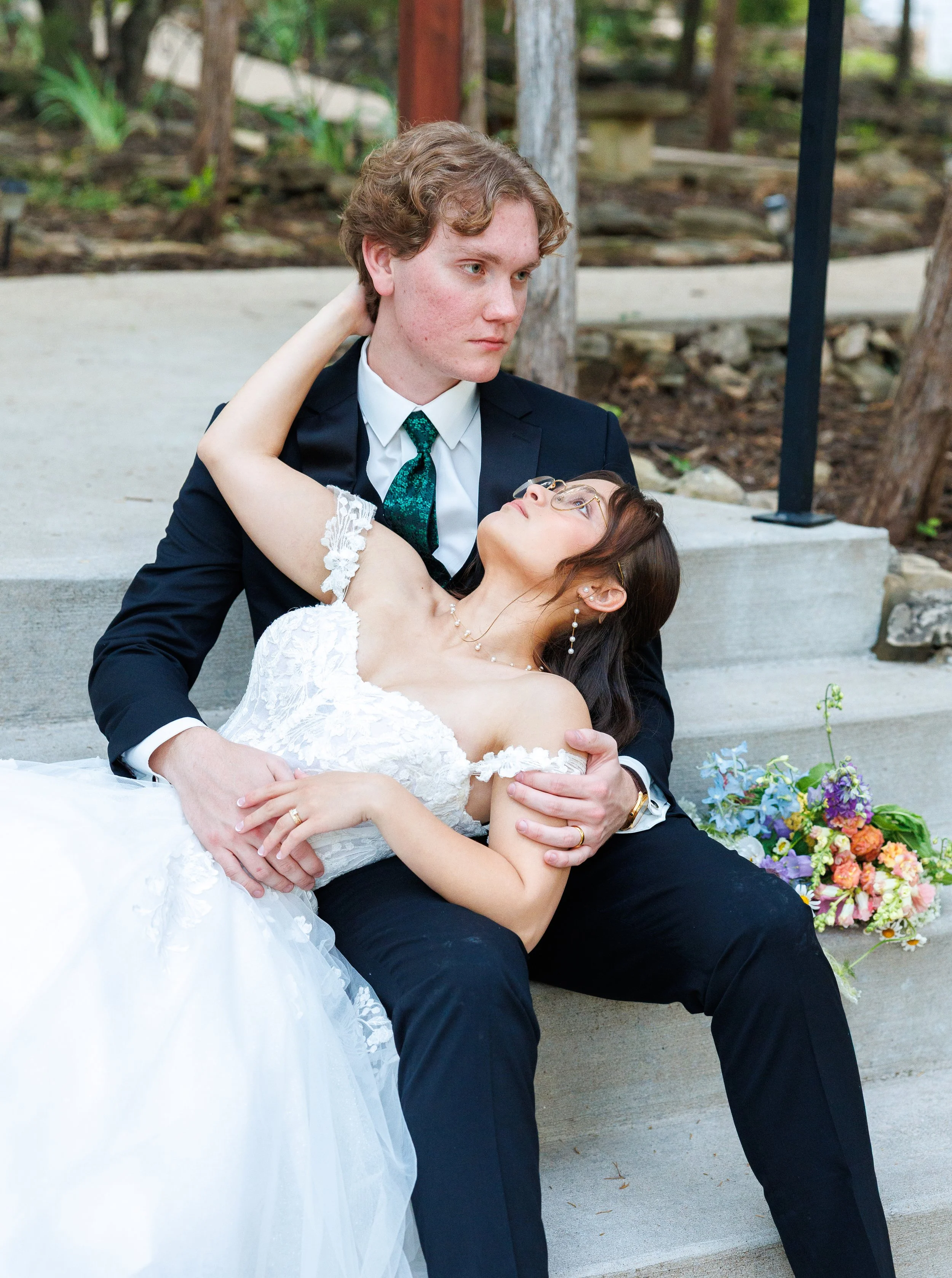 A newlywed couple sitting on concrete stairs outdoors, with the groom holding the bride in a dip, surrounded by flowers and trees.