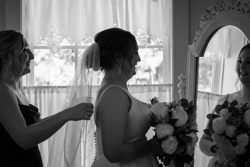 A bride standing in front of a mirror holding a bouquet, smiling, with a woman adjusting her veil in a well-lit room.
