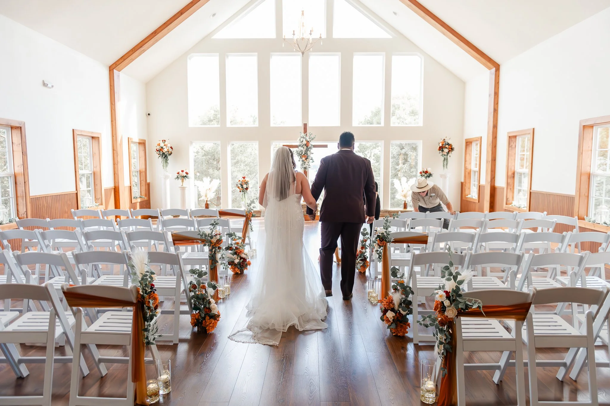 A bride and groom holding hands at the altar in a bright, spacious wedding chapel with large windows, decorated with floral arrangements, before the ceremony begins.
