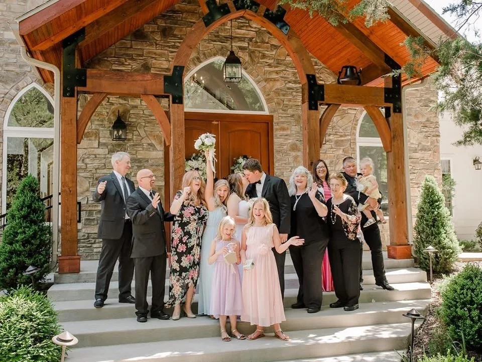 A group of people celebrating outside a house with a stone facade and wooden arch entrance, dressed in formal and semi-formal attire, with some holding drinks and a bouquet, smiling and interacting with each other.