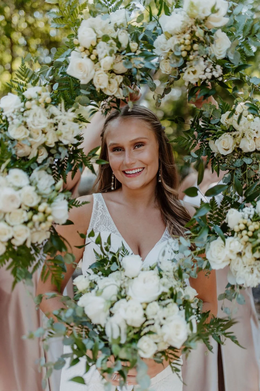 Smiling woman in a white dress holding a bouquet of white flowers, surrounded by a floral arch with white flowers and greenery.