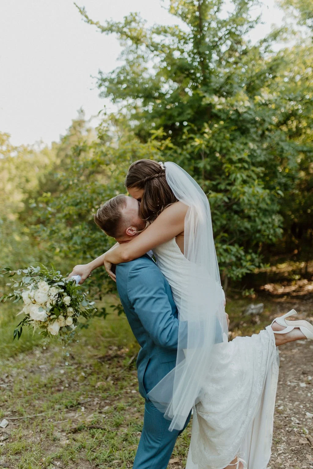A bride in a white wedding dress and veil being lifted by a groom in a blue suit outdoors, with greenery in the background.