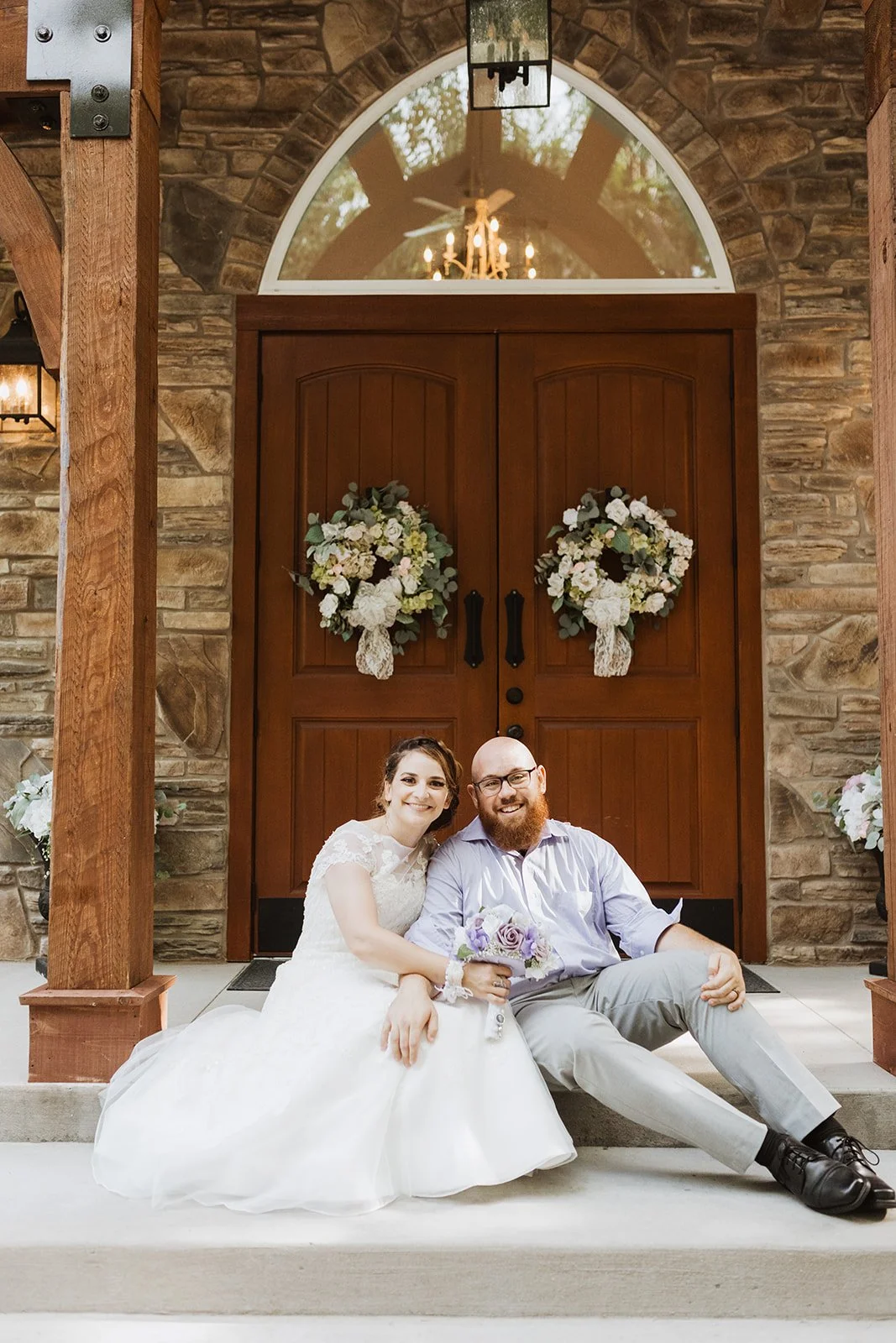 A bride and groom sitting on the steps outside a church entrance decorated with wreaths of flowers on the wooden double doors.