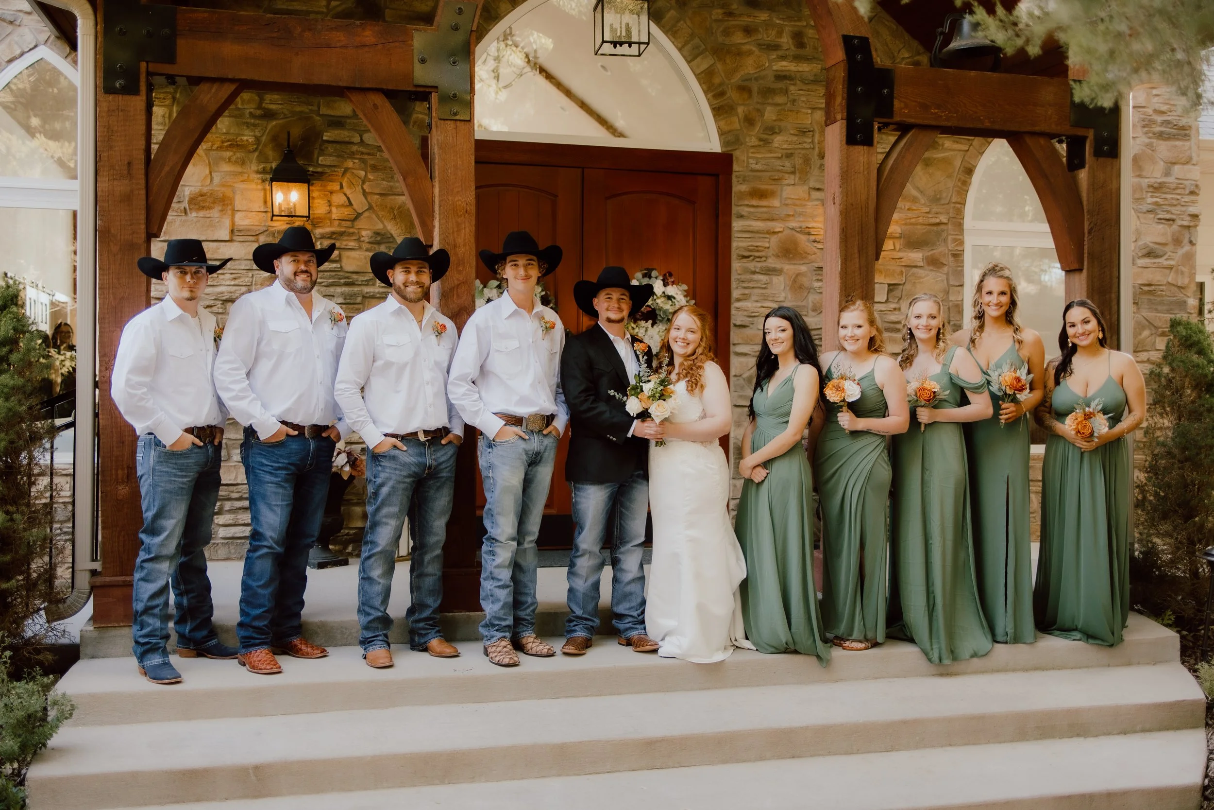 Group of nine people standing on a porch in front of a stone and wooden house, dressed in cowboy and wedding attire, celebrating a wedding.