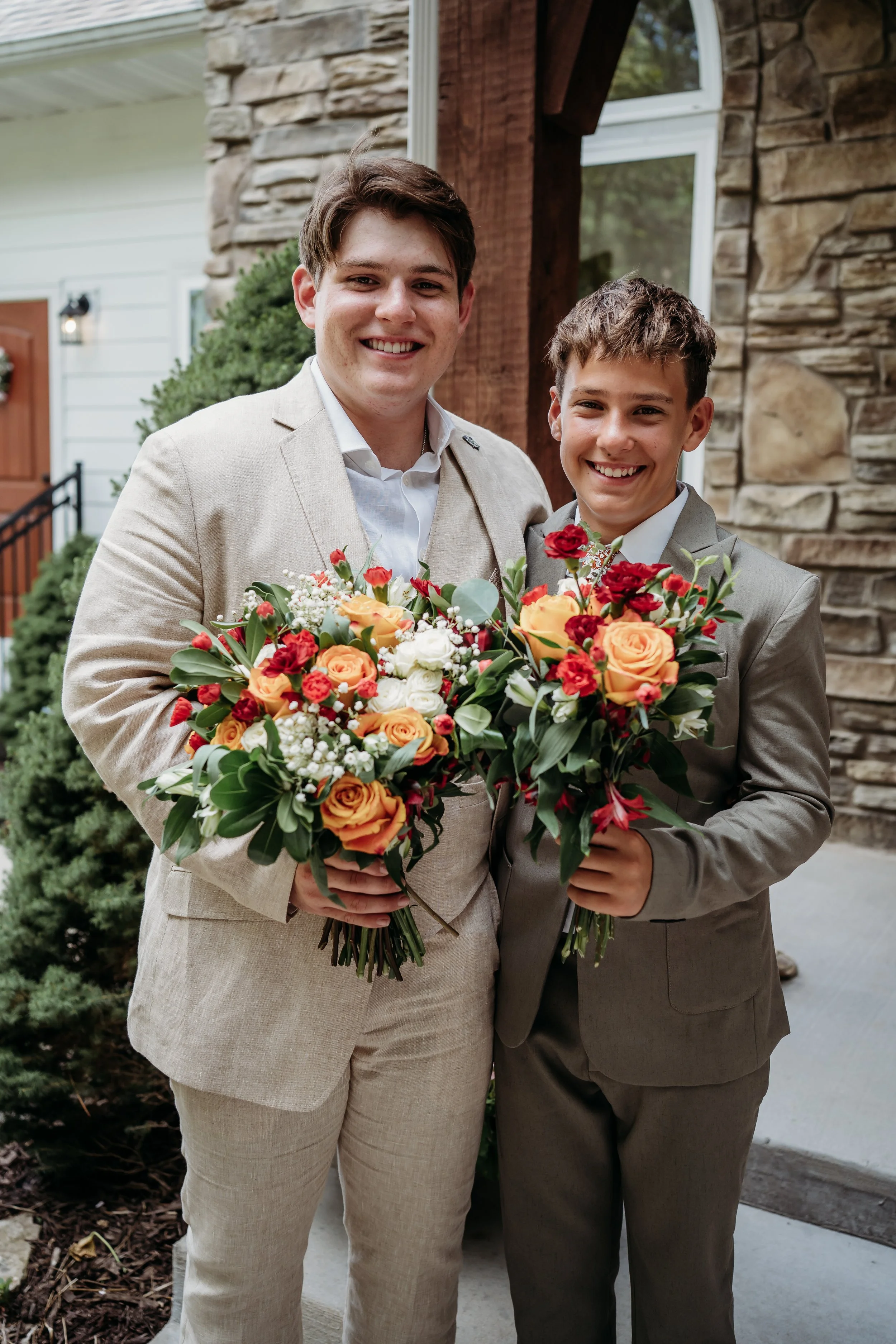 Two young men in suits smiling and holding a bouquet of colorful flowers outside a house.