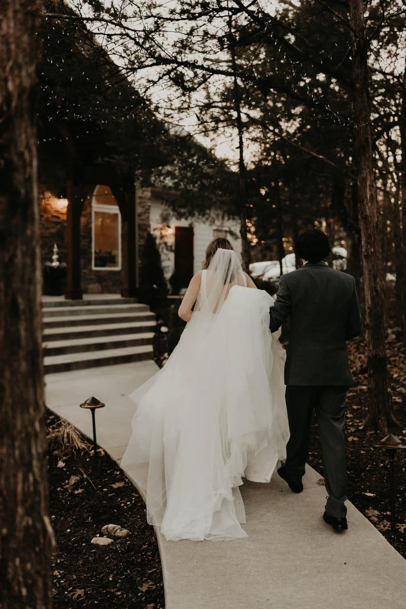 A bride and groom walking outside on a wedding day during dusk, surrounded by trees and in front of a house with stairs.