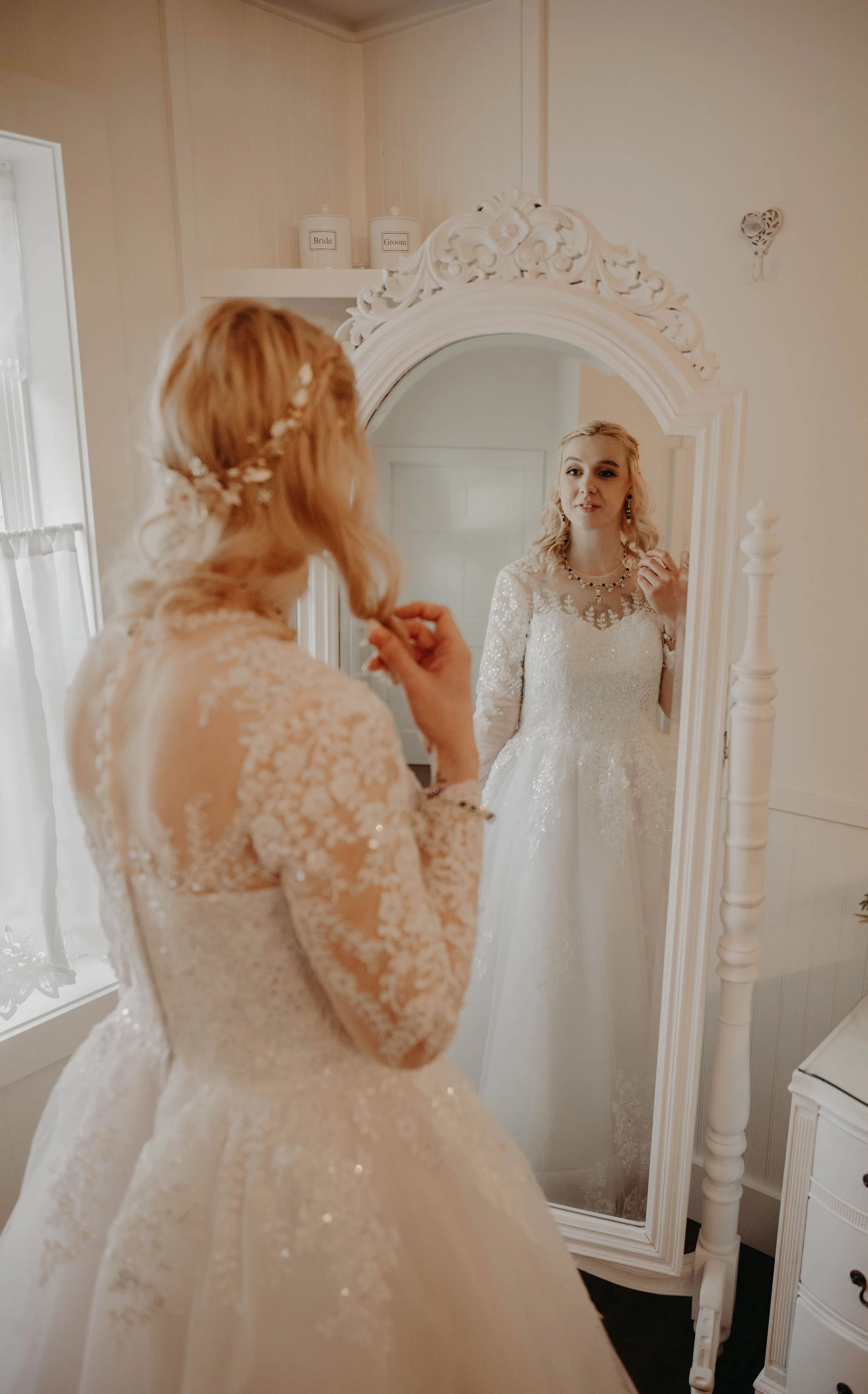 A woman with blonde hair in a lace wedding dress looks at herself in a large ornate mirror, holding a small object in her hand.
