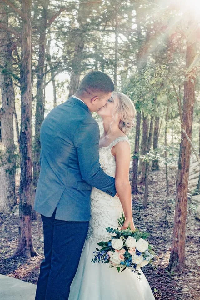 A newlywed couple sharing a kiss in a forested area, with the bride holding a bouquet of white and pink flowers and the groom wearing a blue suit.