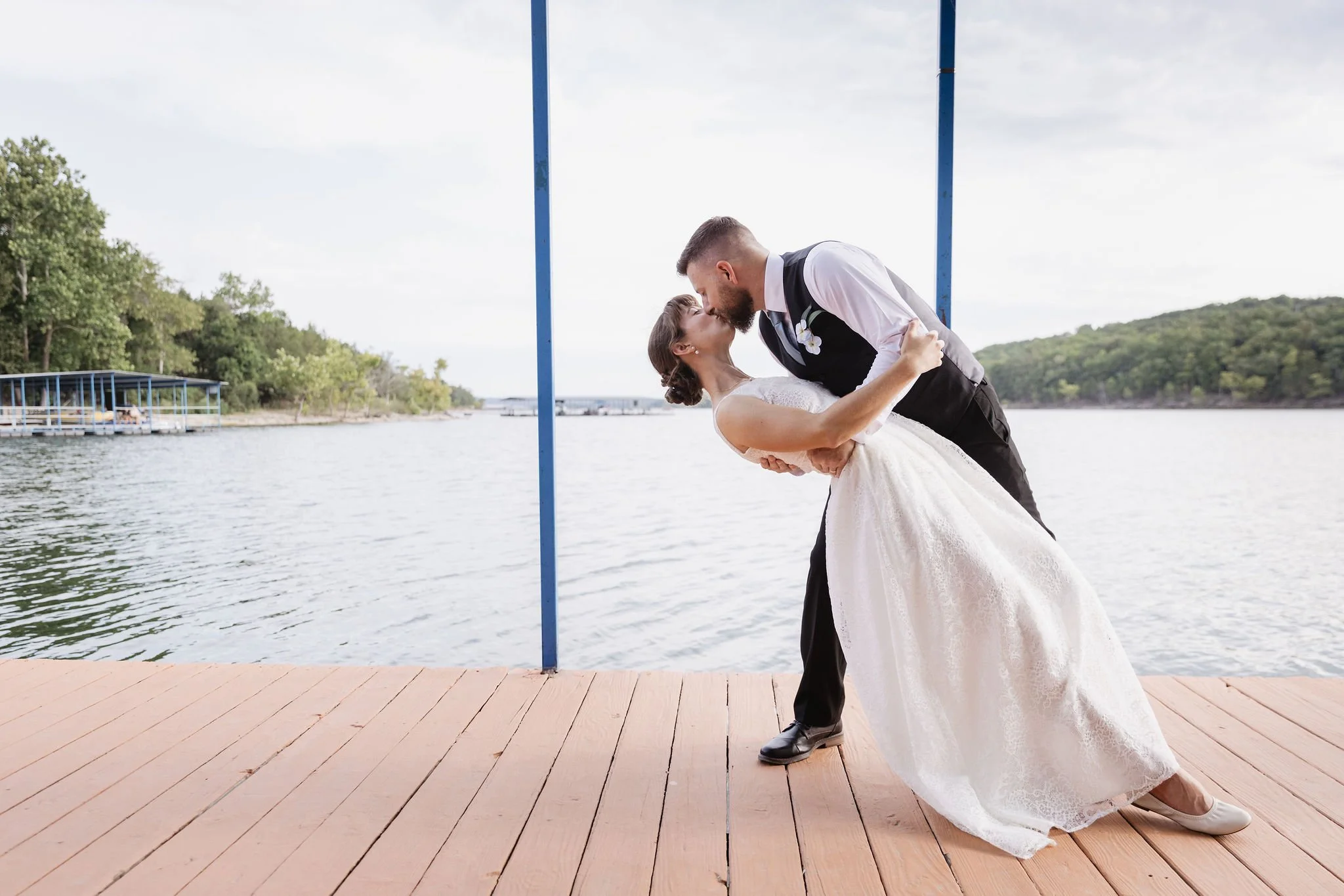 A newlywed couple sharing a kiss by a lakeside, with the bride in a white wedding gown and the groom in a black suit and vest, on a wooden dock with trees and water in the background.