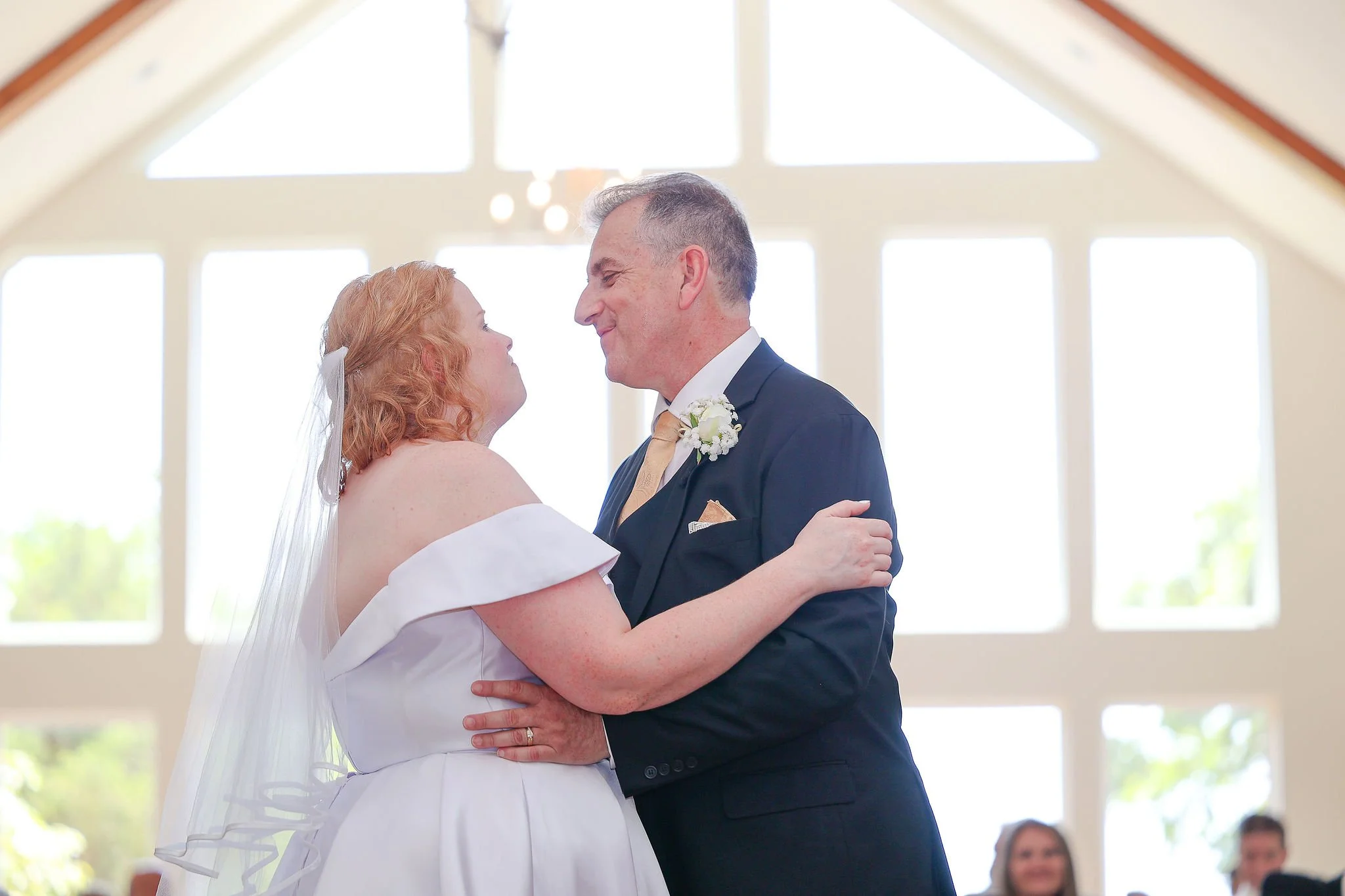 A bride and groom dancing during their wedding ceremony inside a bright hall with large windows, with guests watching in the background.