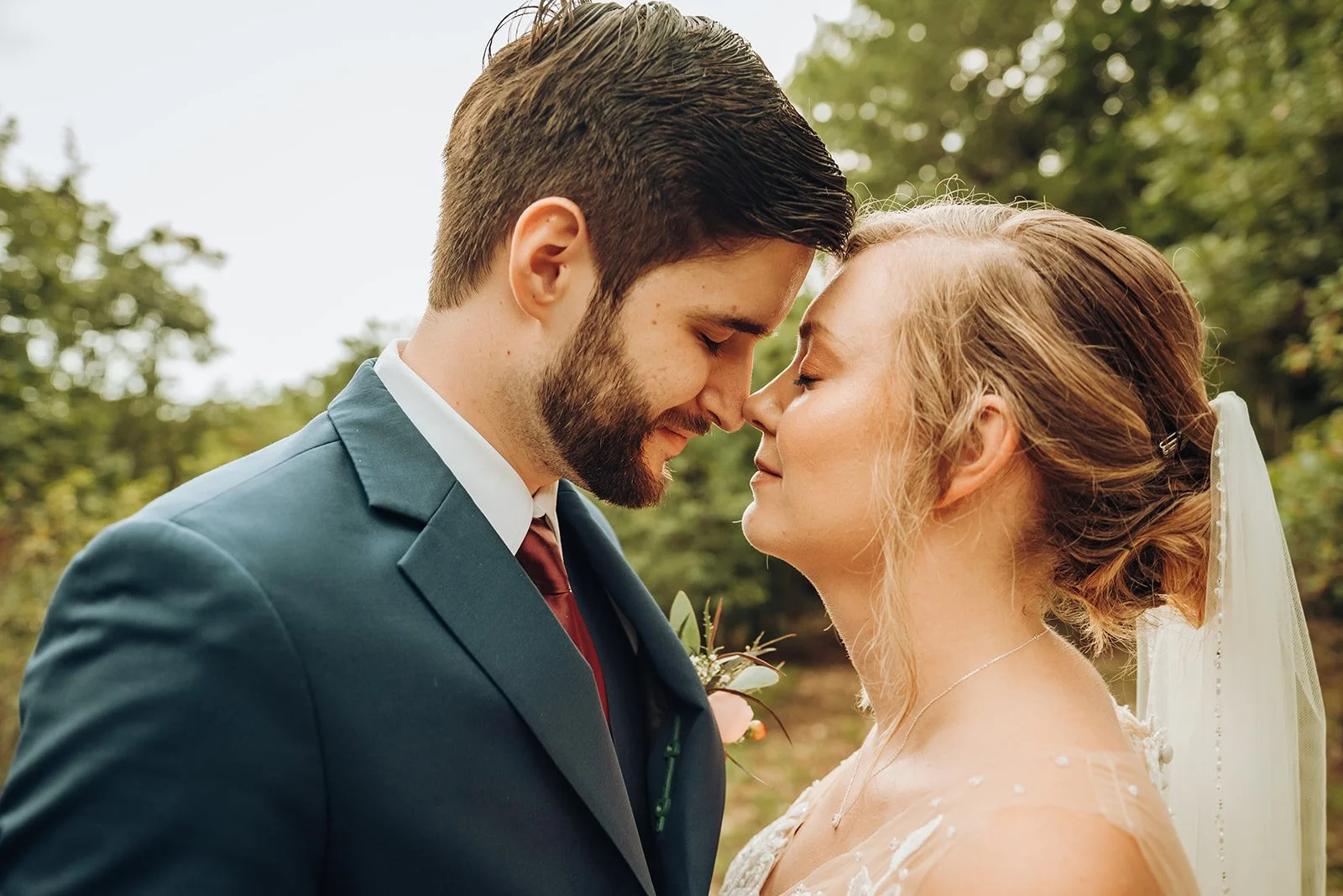 A bride and groom with their foreheads touching outdoors during a wedding, with trees in the background.