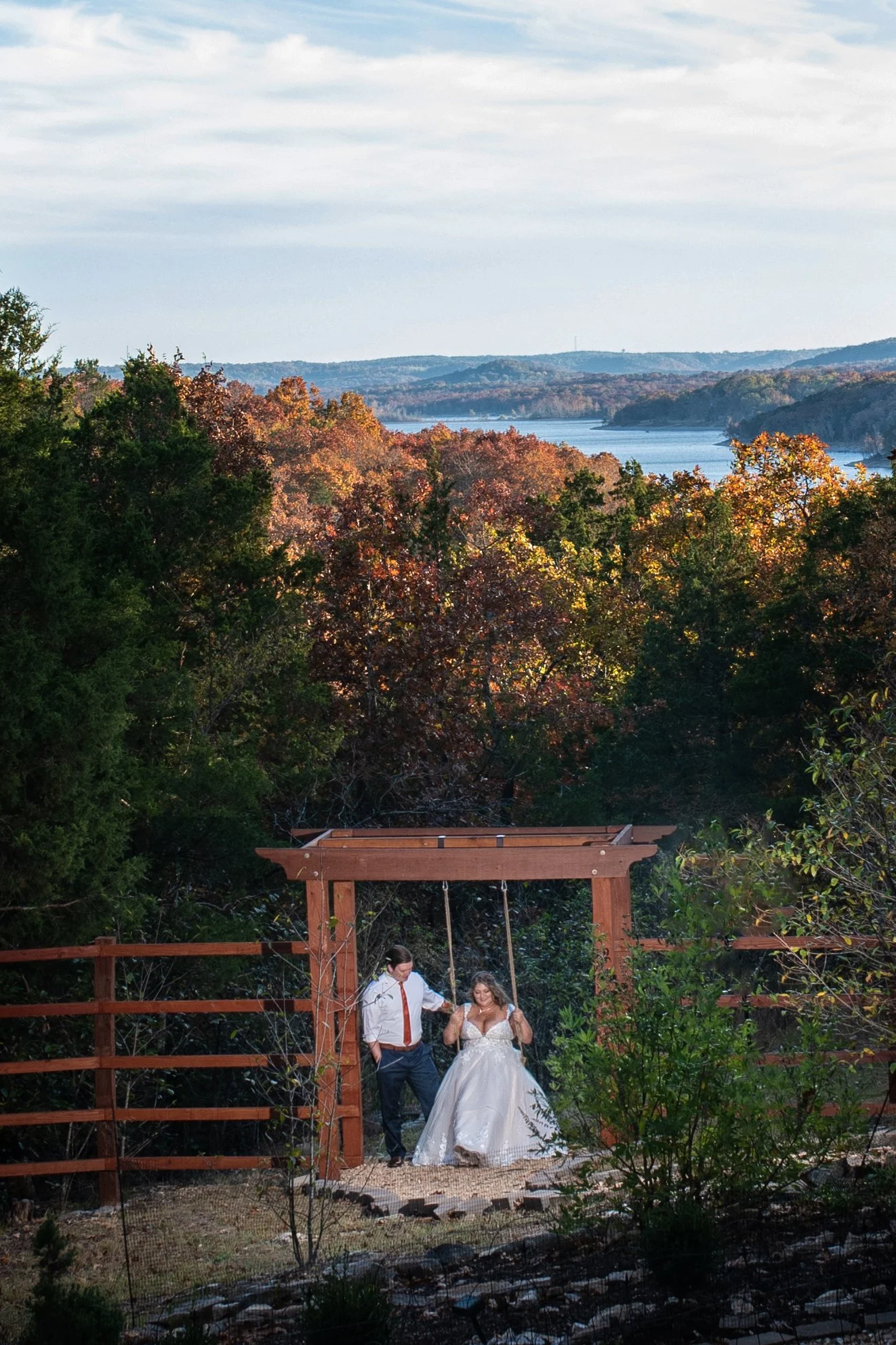 A couple on a swing set in a backyard with lush trees, a lake, and rolling hills in the background, during the daytime.
