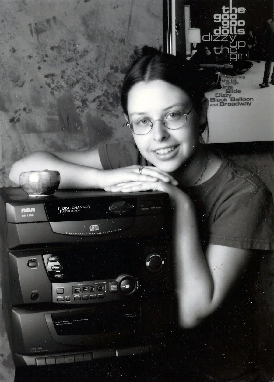 One of Erin Dorney’s black and white senior pictures from high school, showing a person with glasses smiling and hugging a gigantic 5-disc CD stereo with a poster of the Goo Goo Dolls in the background