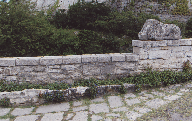 Photograph of a stone lion, weathered by the elements, at the end of long stone wall