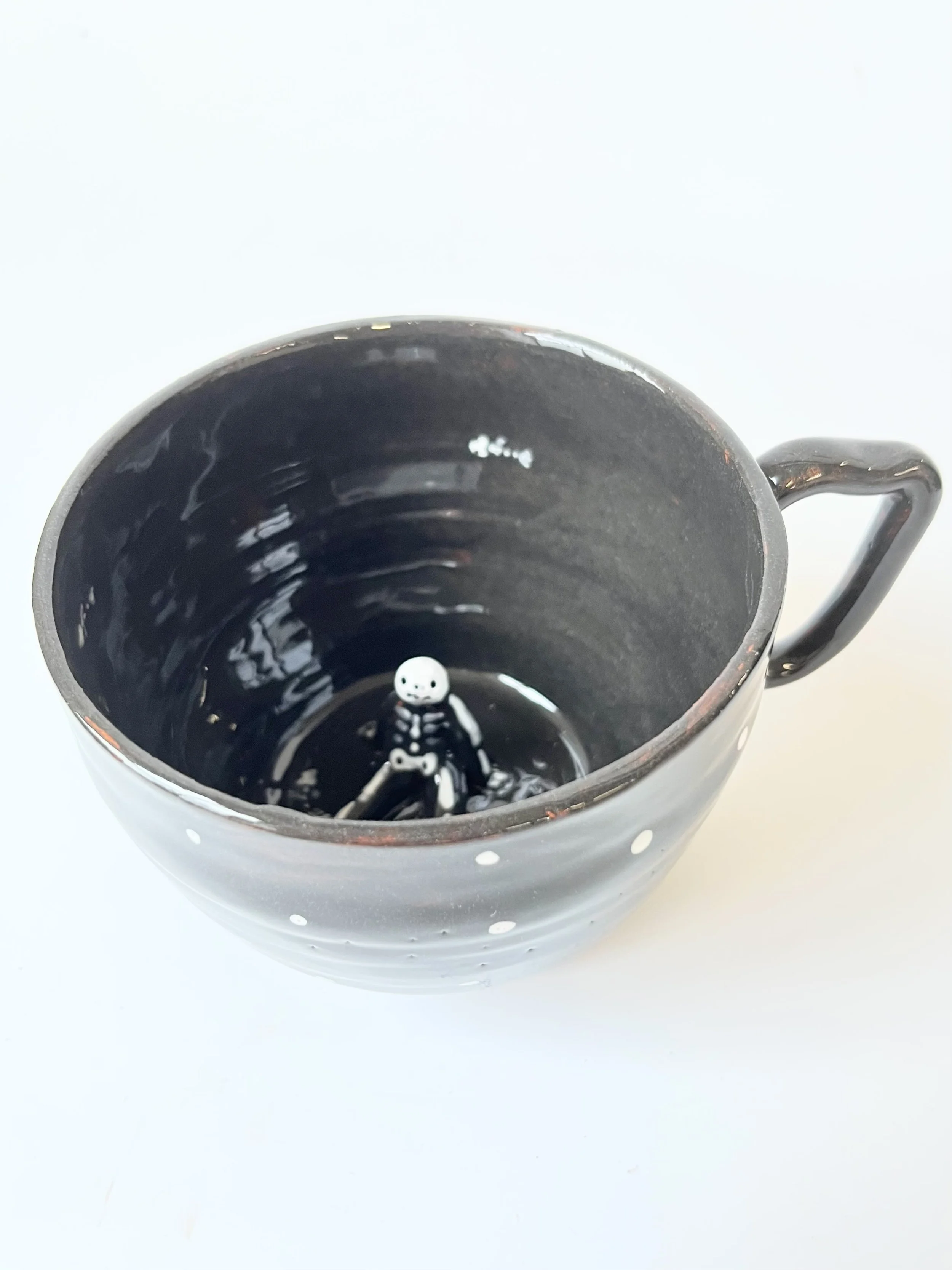 Black earthenware cup with tiny skeleton sitting on the bottom  and a very fancy handle