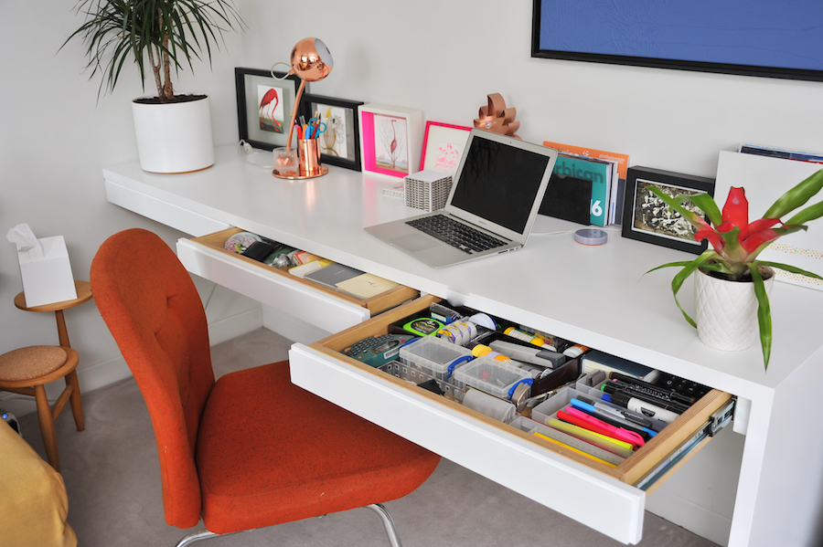 A red chair sits against a clean, bright white home office, where the modular desk has multiple drawers pulled out to show the detailed and efficient organisation inside.