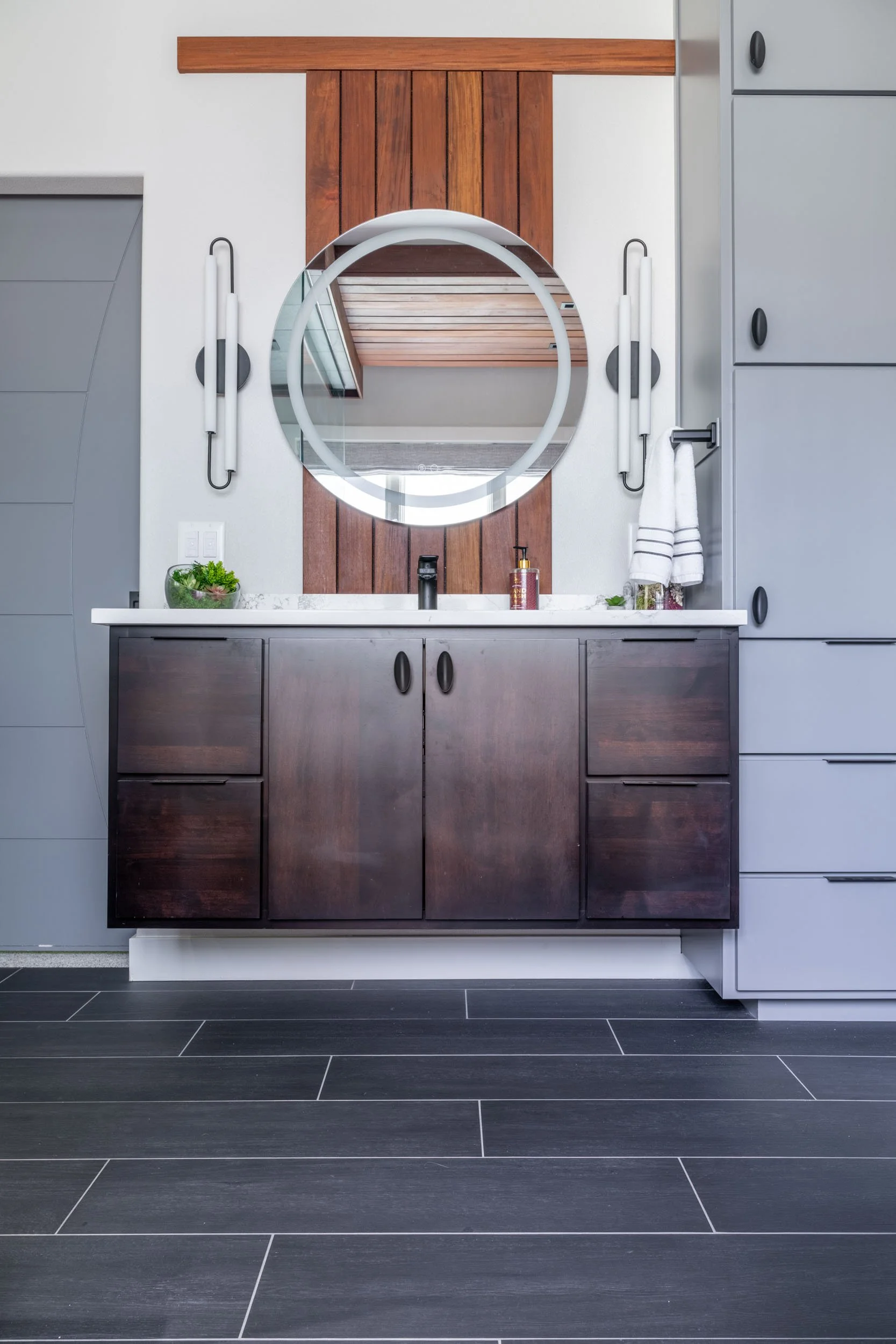 A focused view of the vanity featuring the lighting, tall cabinetry, and teak accents behind the mirror.