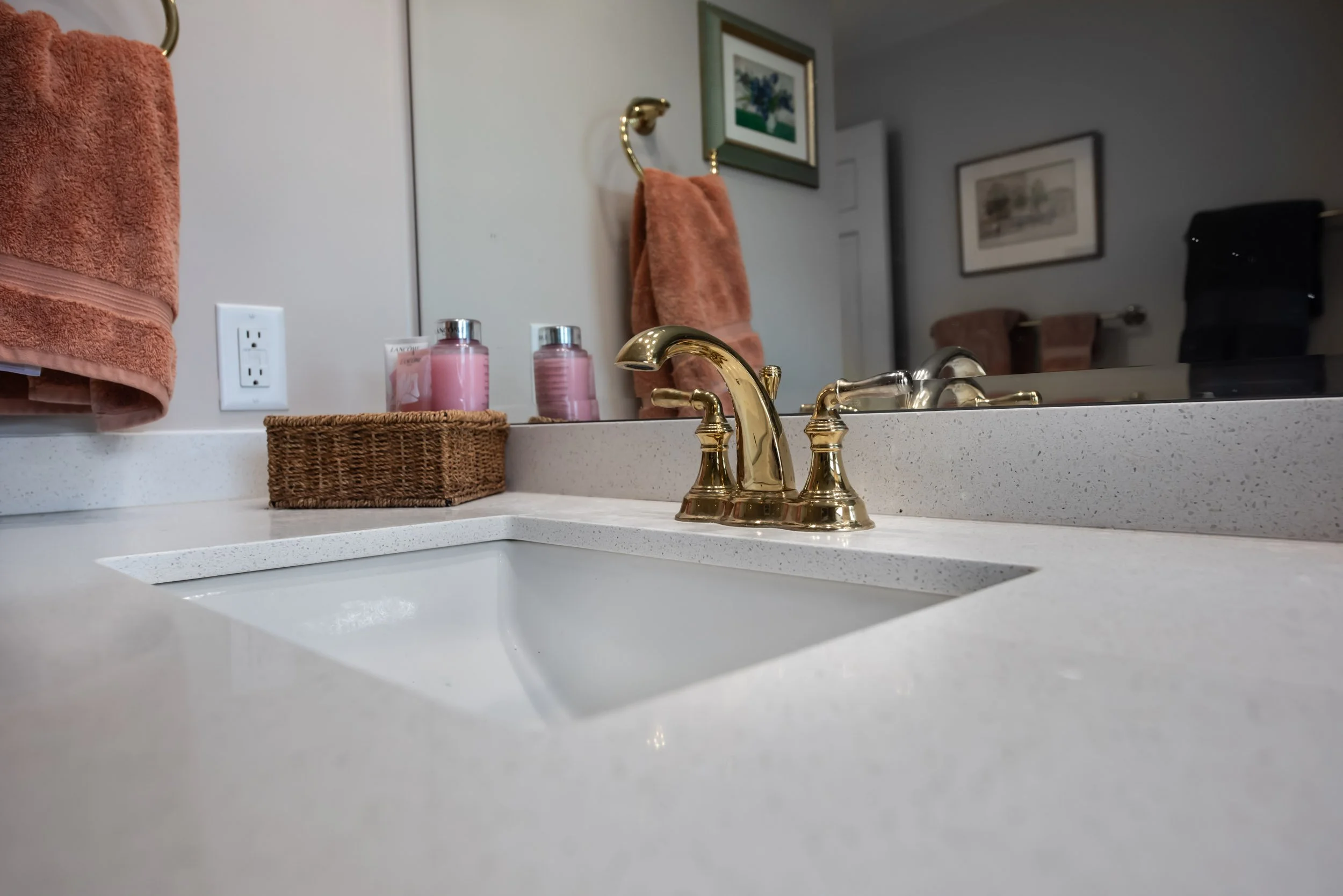 A light colored quartz vanity tope with gold fixtures and a frameless mirror