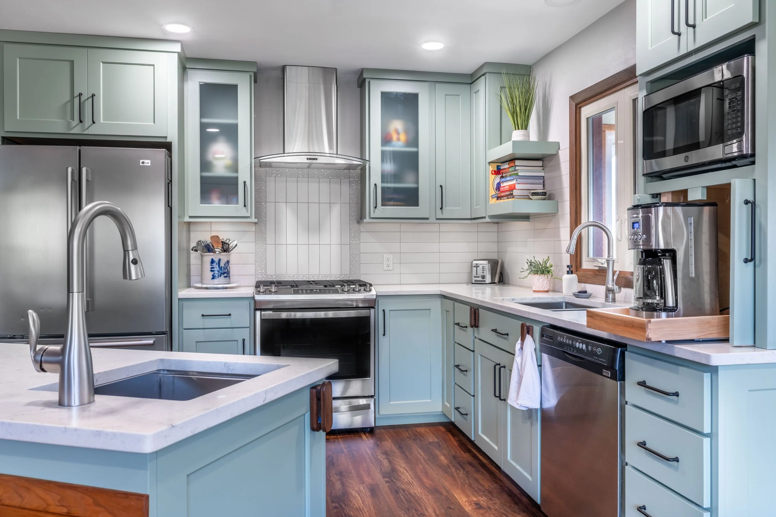 A L-shaped kitchen with an island and prep sink with light green cabinets, grey striated quartz countertops, and stainless steel appliances