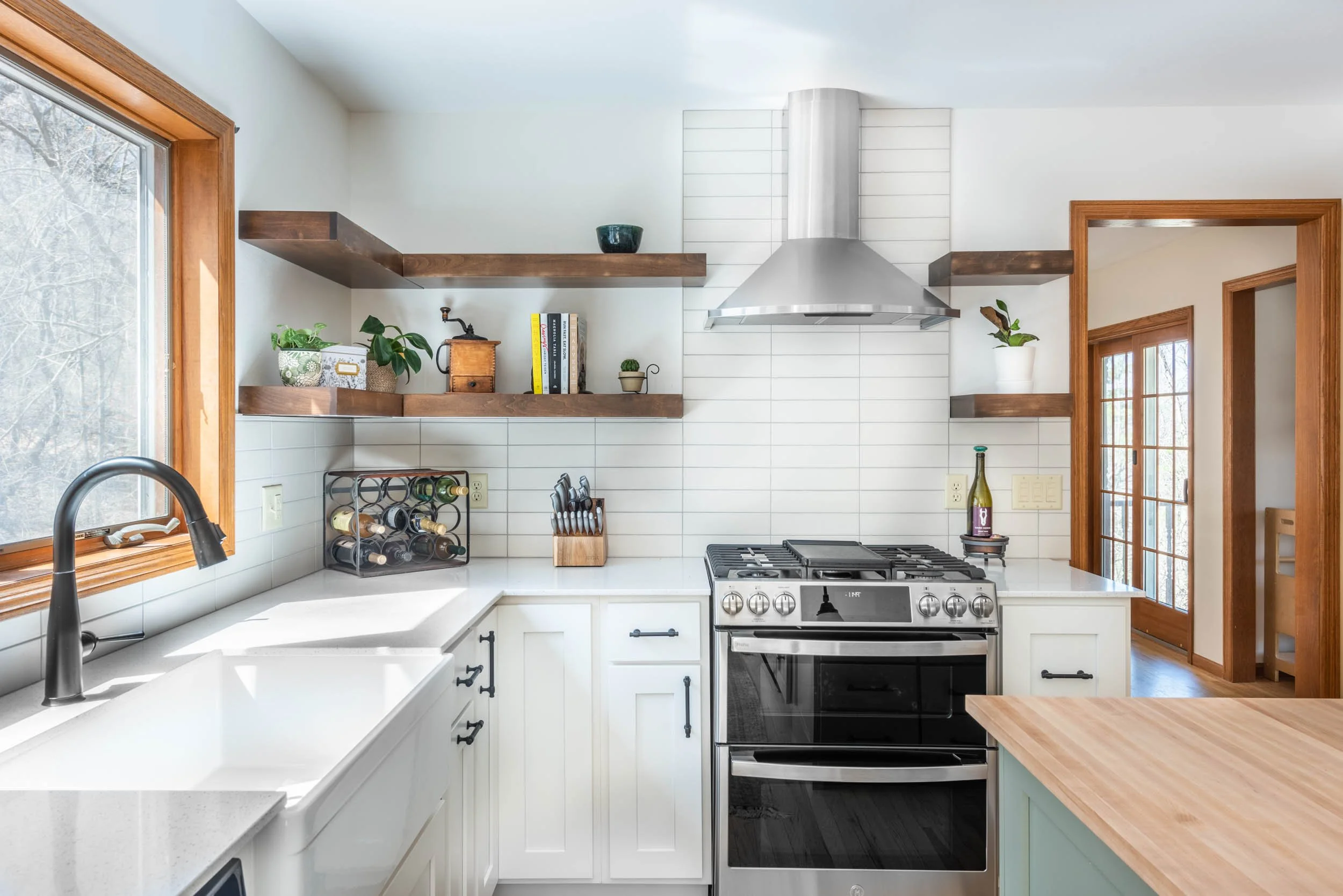 This kitchen uses white cabinets and white backsplash tile, accented by a light green island cabinet with butcherblock countertop and dark stained open shelving.