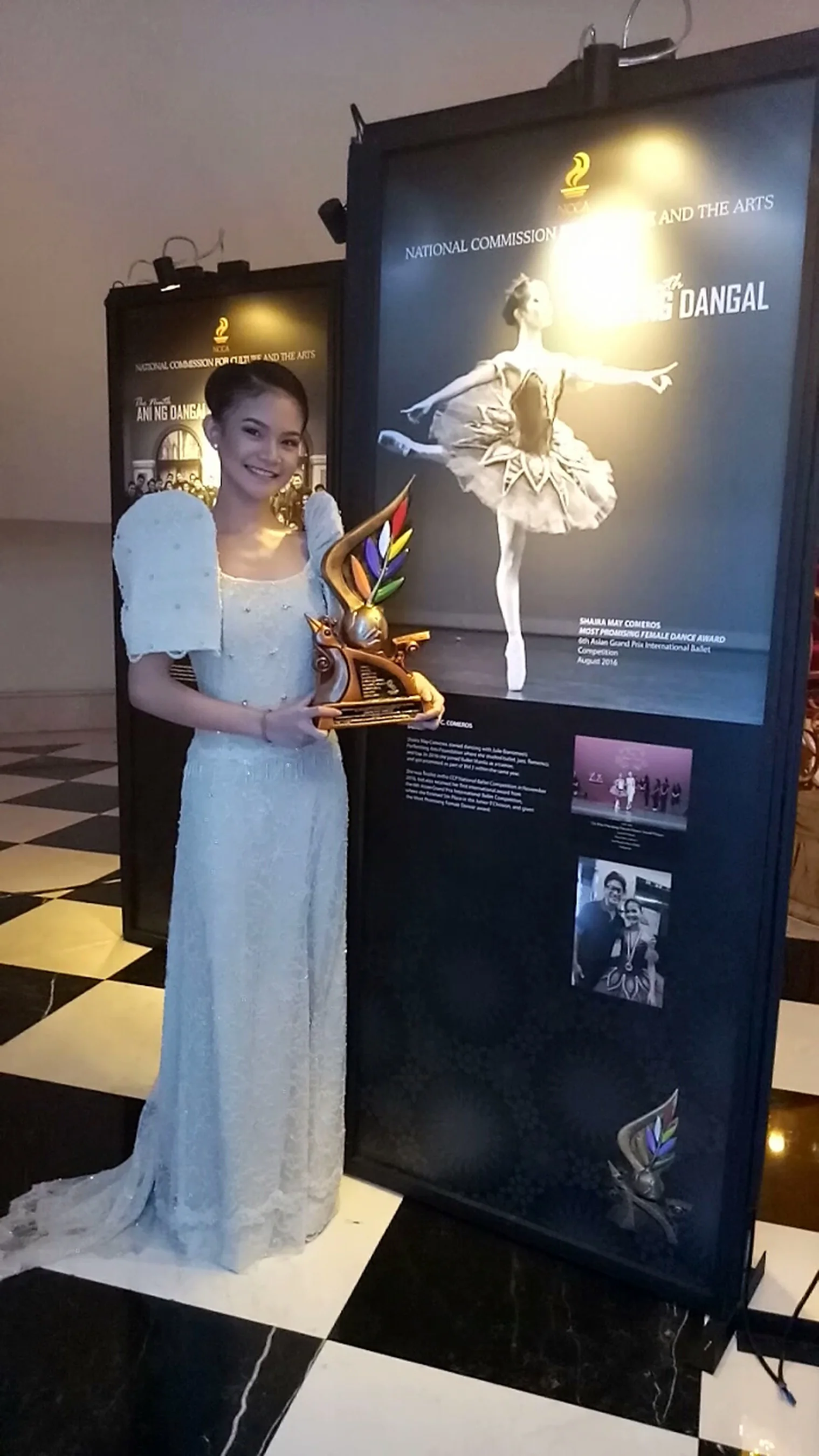Carrying her Sarimanok trophy, Shaira Comeros stands beside a panel showcasing her achievements in dance.