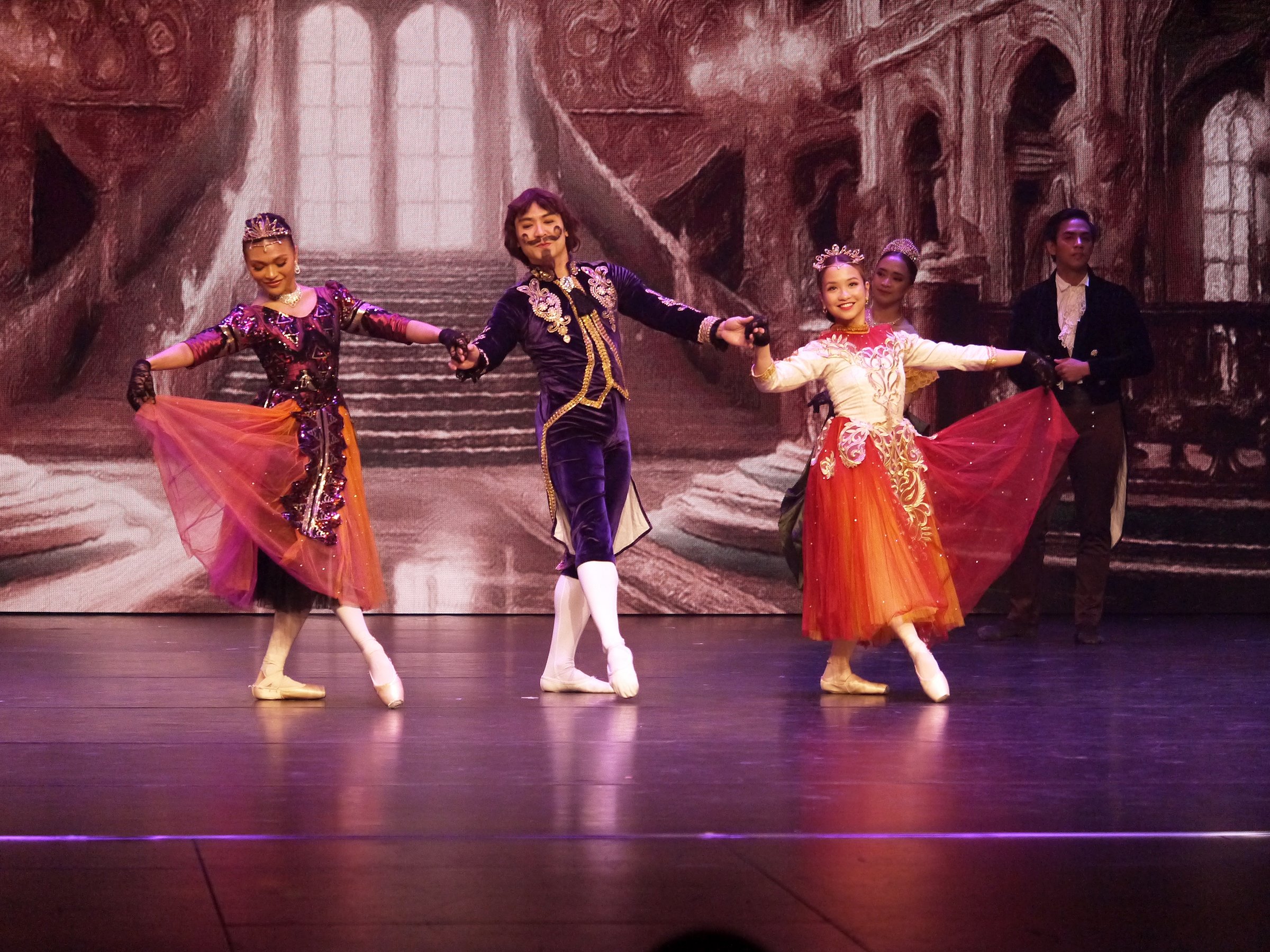    Dapper in his purple tailcoat, the Duke (Romeo Peralta Jr.) escorts two young princesses played by Celine Astrologo (left) and Jasmine Pia Dames as they arrive at the ball in Lisa Macuja Elizalde’s  Cinderella  (2025). The fairy-tale ballet was br