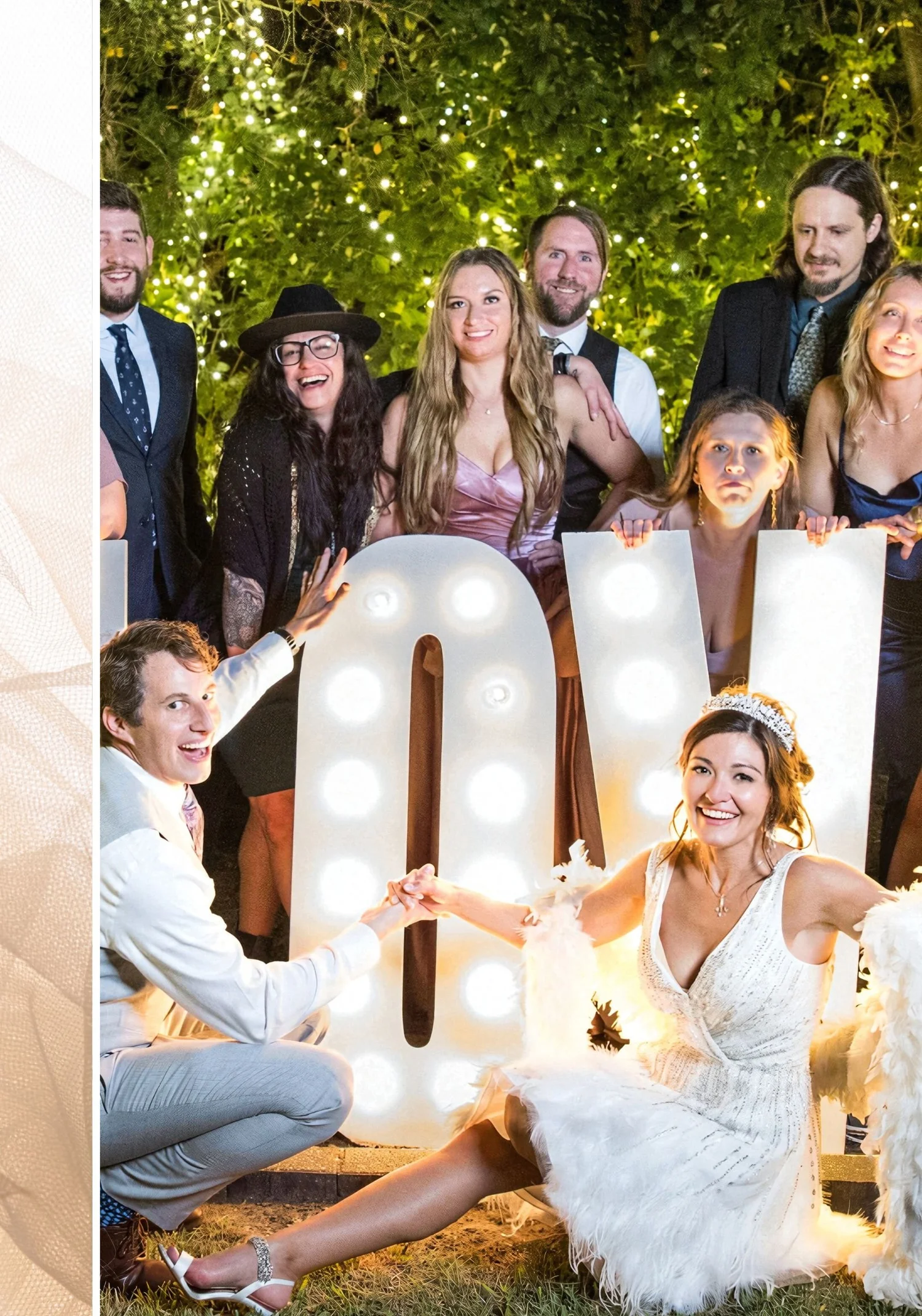 A bride in a short beaded dress and tiara sitting before glowing LOVE marquee letters at night, the groom and their wedding guests gathered behind them