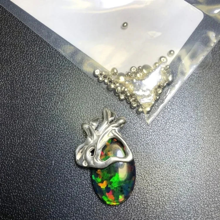 An organic cast silver form set over a cabochon flashing green, red and blue, beside a bag of small cast droplets